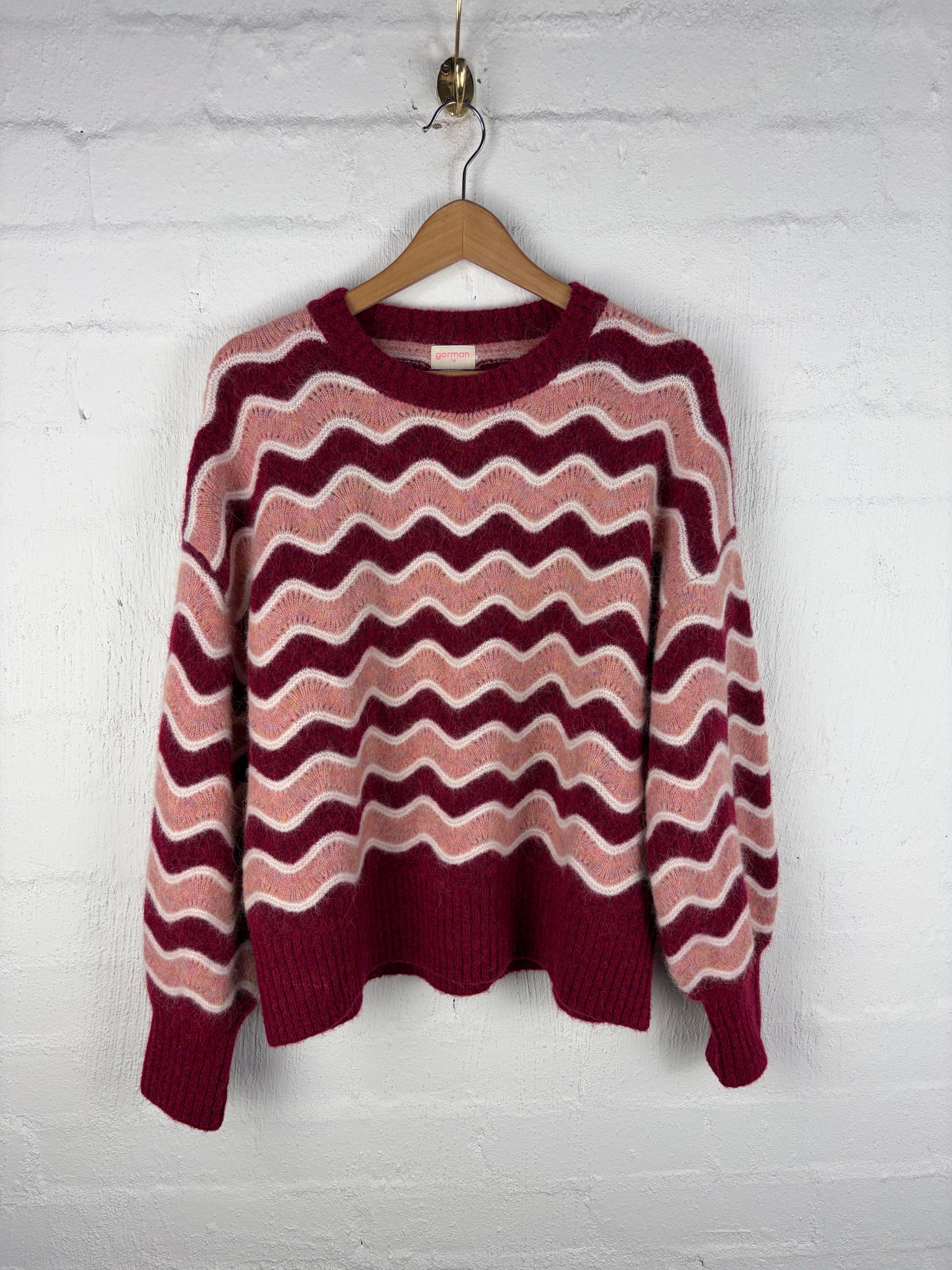 A knit sweater with a wavy chevron pattern in burgundy, dusty pink, and cream, hanging on a wooden hanger against a white painted brick wall.