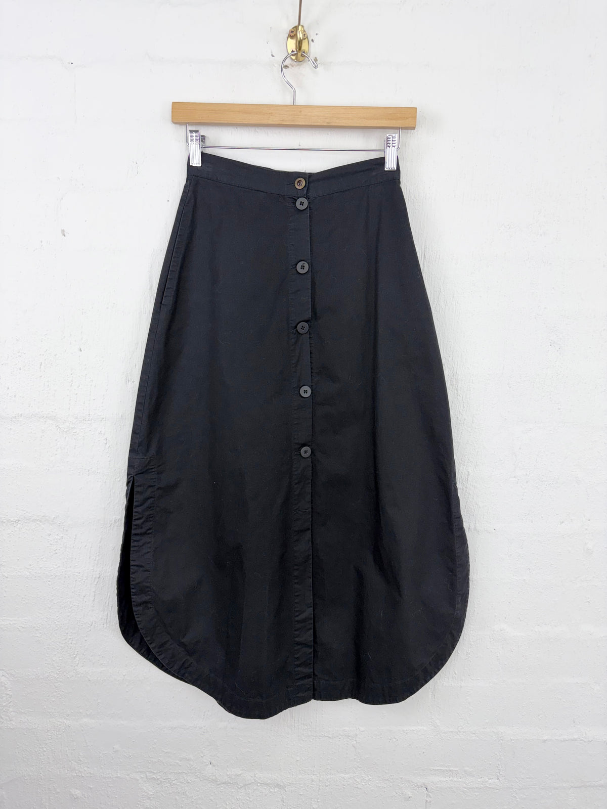 A black midi skirt with a button-down front, hanging from a wooden hanger against a white painted brick wall.