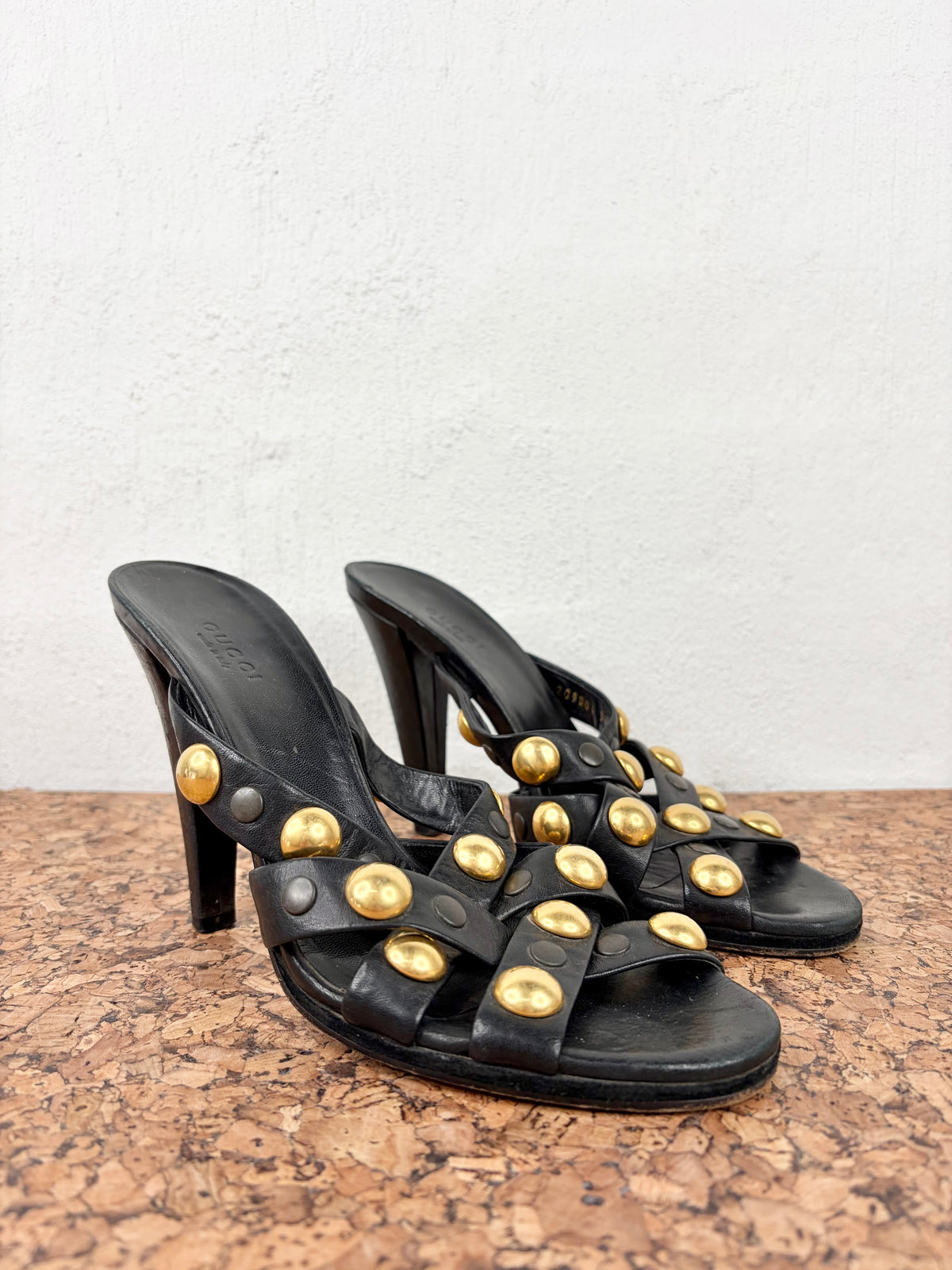 A pair of black leather high-heeled mule sandals with wide straps adorned with large gold-tone studs, displayed on a cork surface against a white wall.