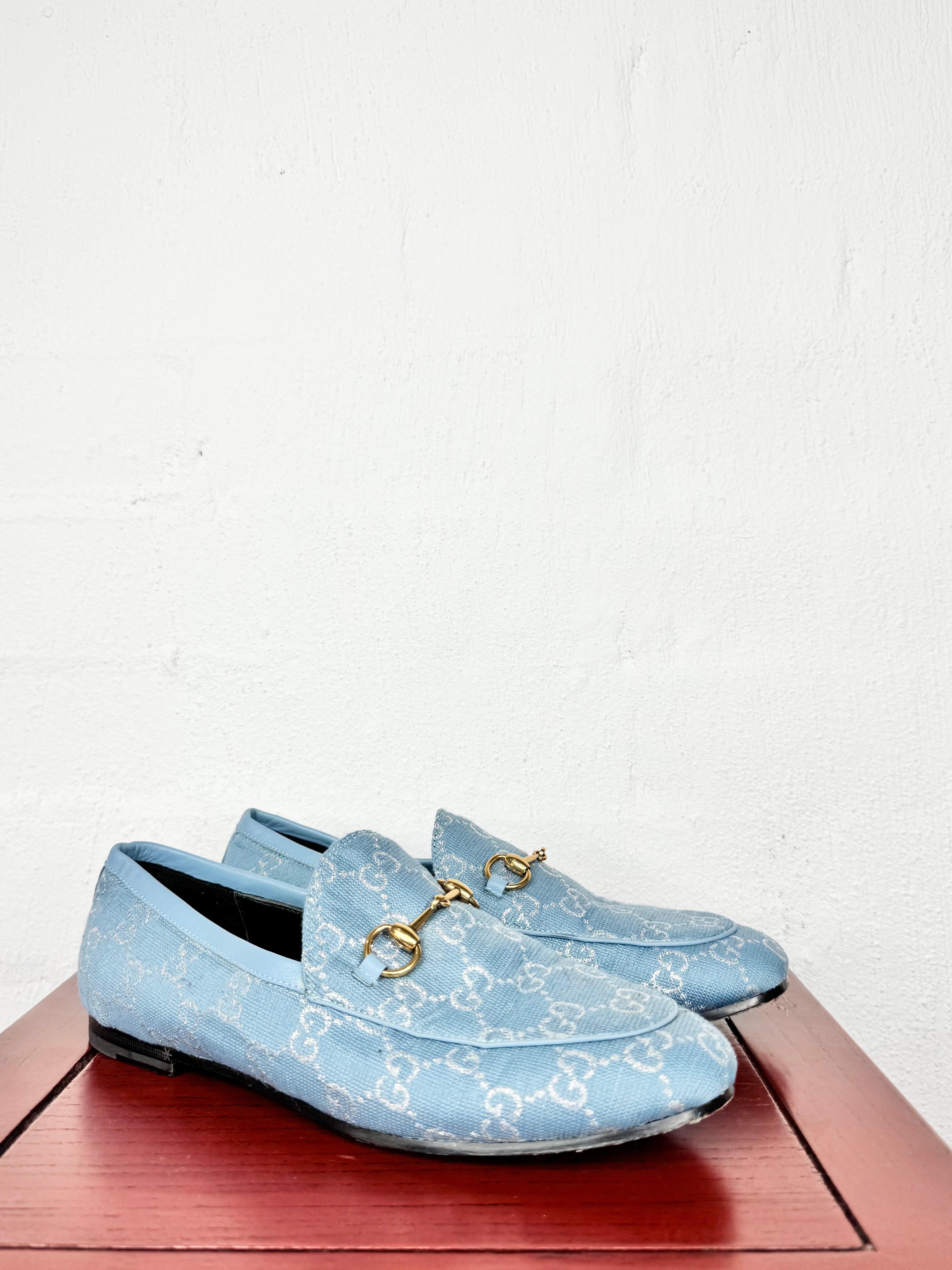 A pair of light blue Gucci loafers with a white GG monogram pattern and gold horsebit details, displayed on a reddish-brown wooden surface against a white wall.