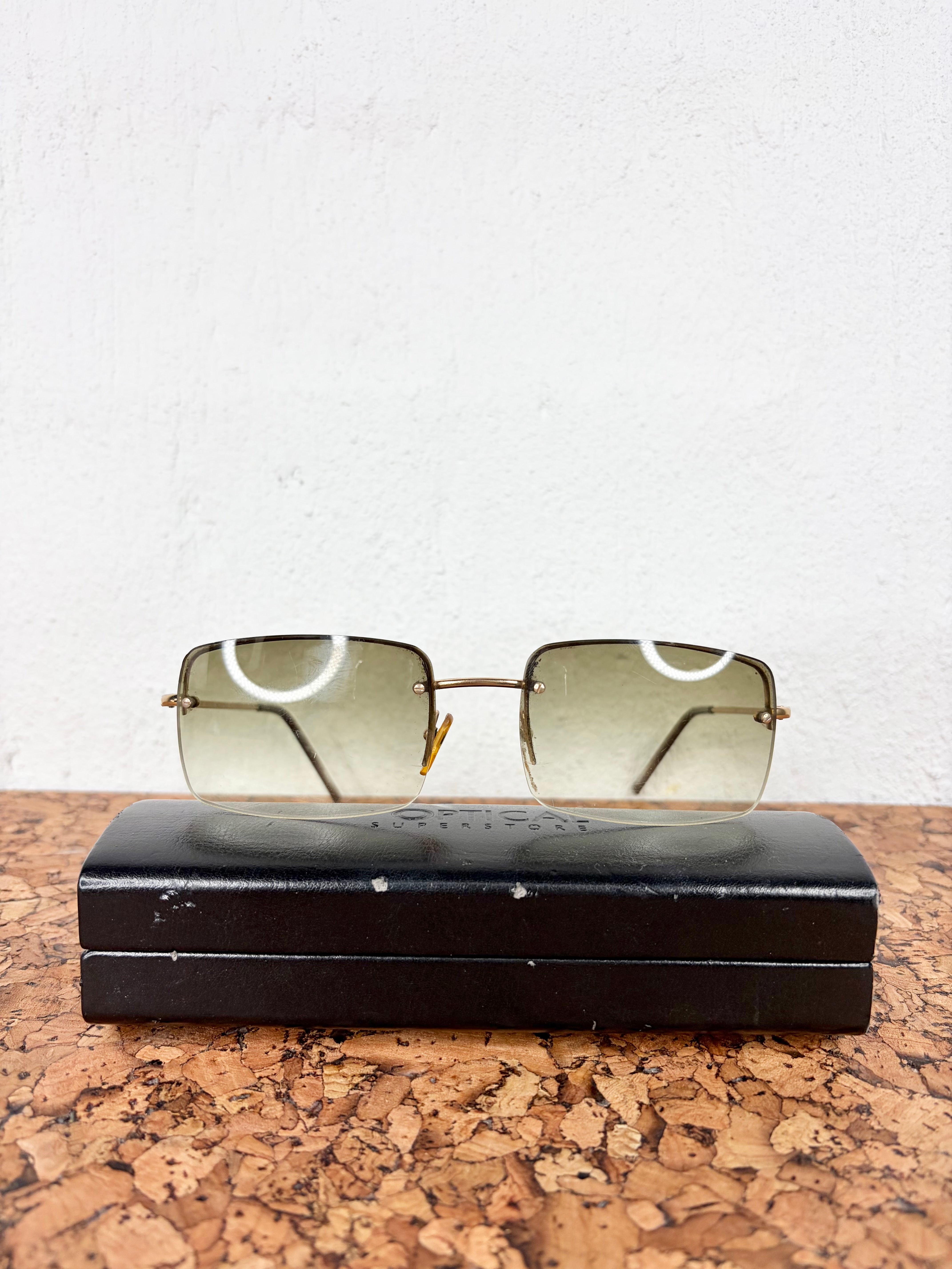 Square rimless sunglasses with gold metal frames and tinted lenses, resting on a black hard case over a cork surface against a white wall.