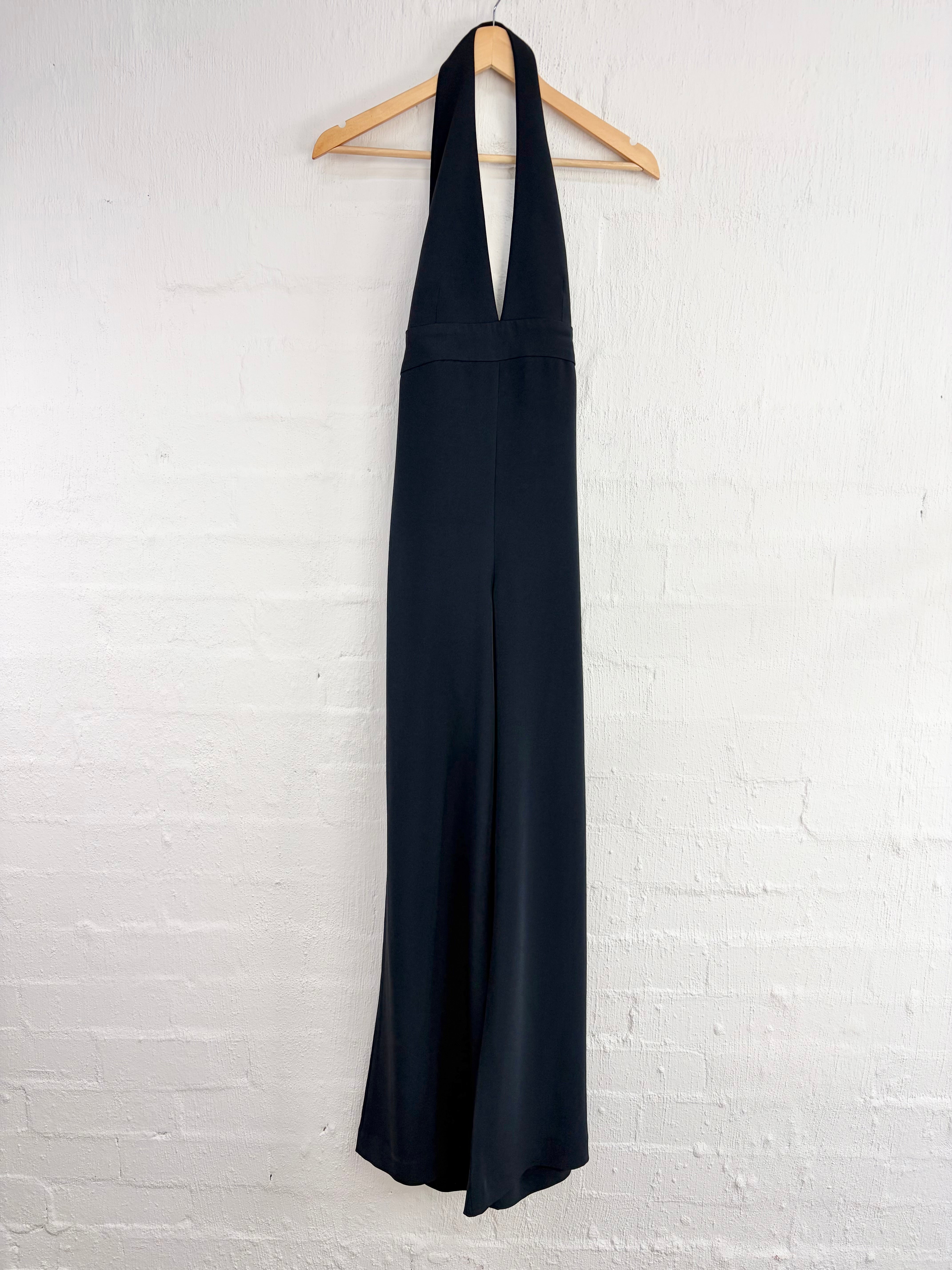 A black halter-neck maxi dress hanging on a wooden hanger against a white painted brick wall.