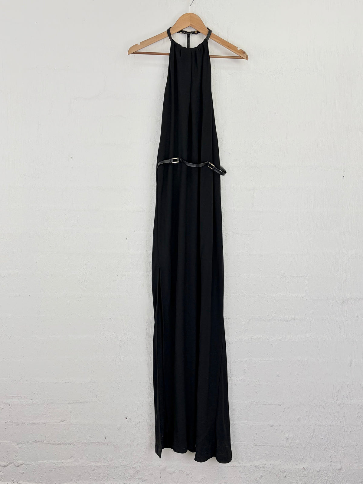A black halter-neck maxi dress with a thin black belt at the waist, hanging on a wooden hanger against a white painted brick wall.