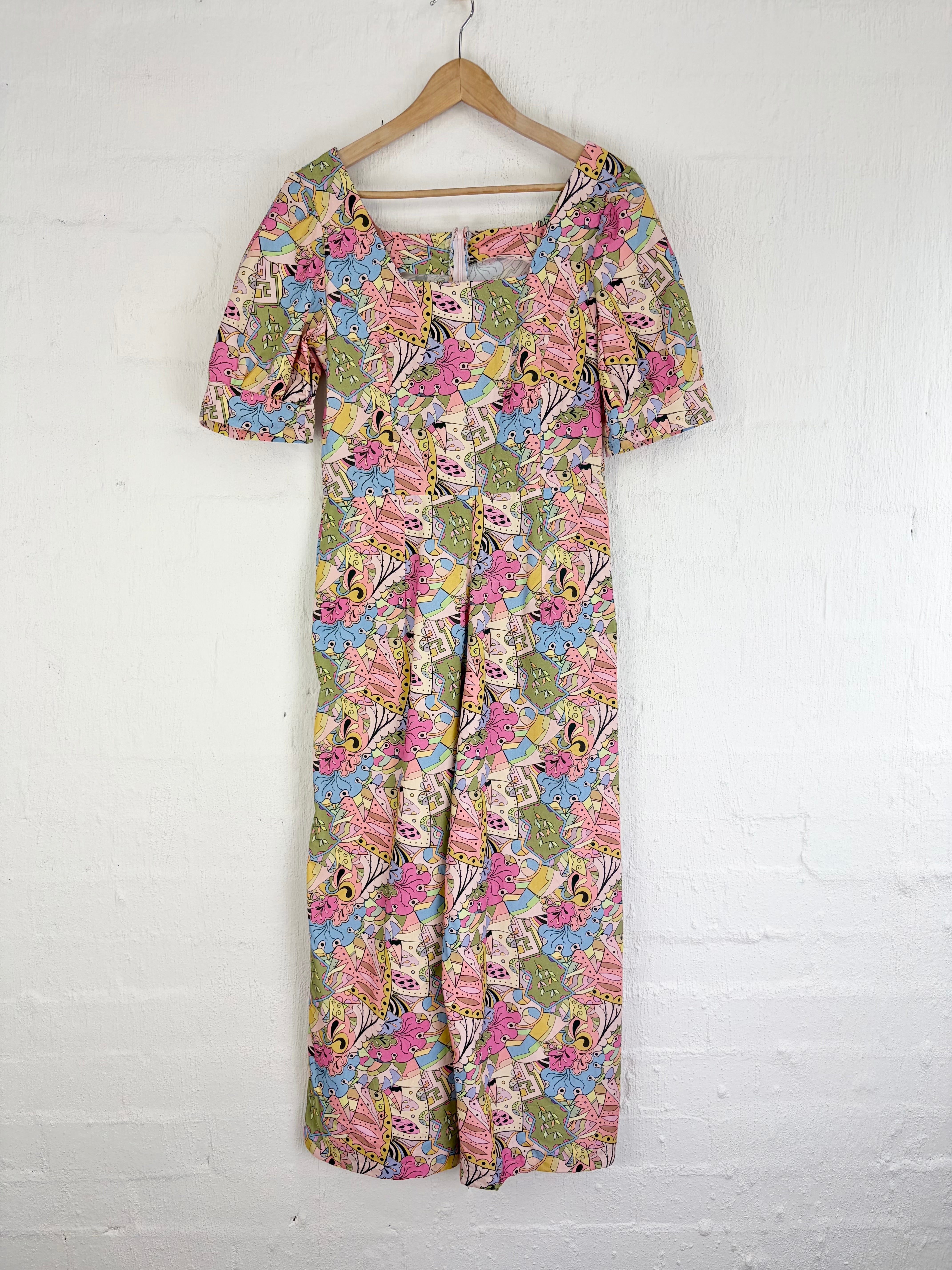 A short-sleeve maxi dress with a colorful floral and abstract print, hanging on a wooden hanger against a white painted brick wall.