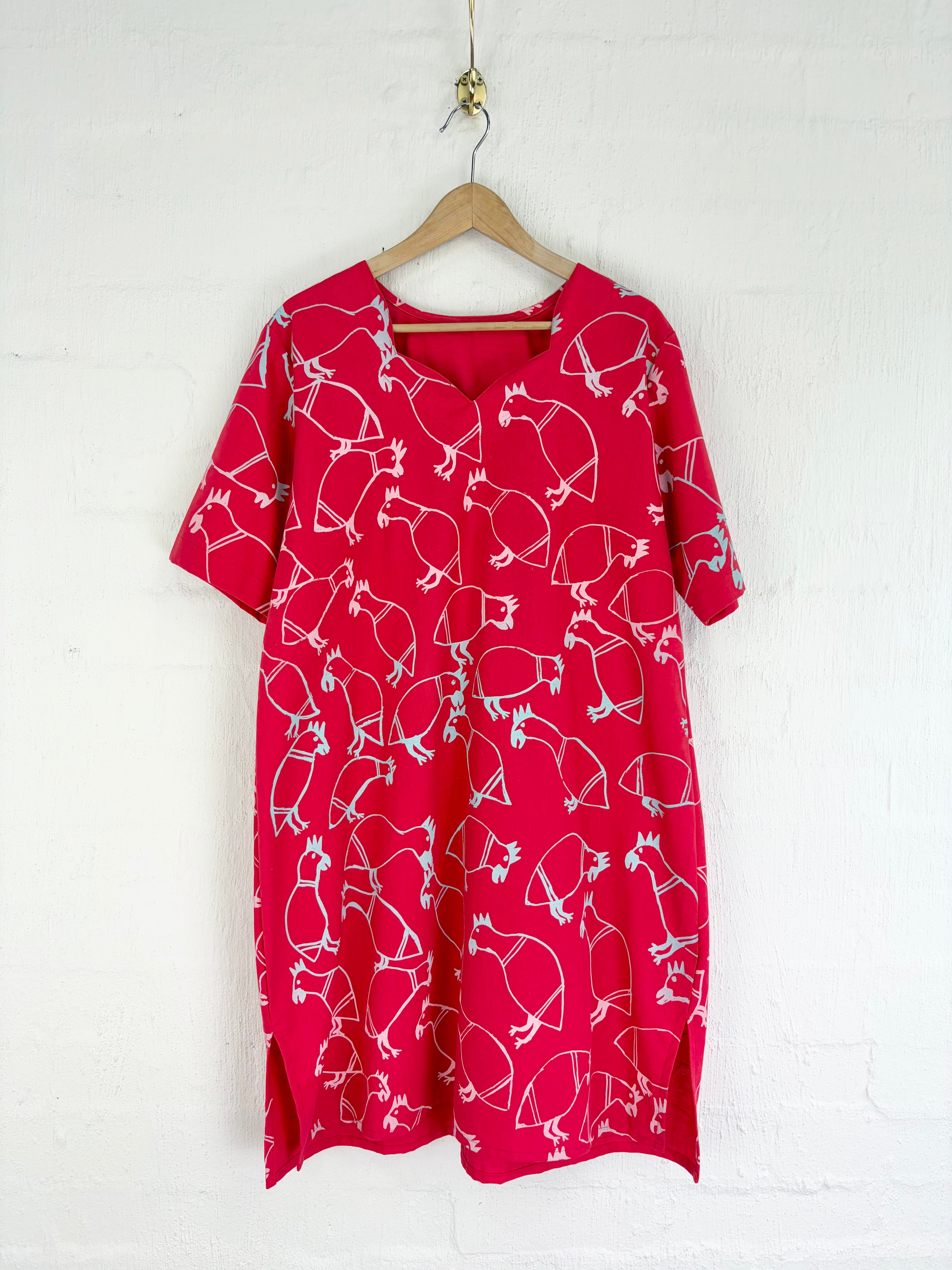 A red short-sleeve dress with a white line-art pattern of bows and arrows, hanging on a wooden hanger against a white brick wall.