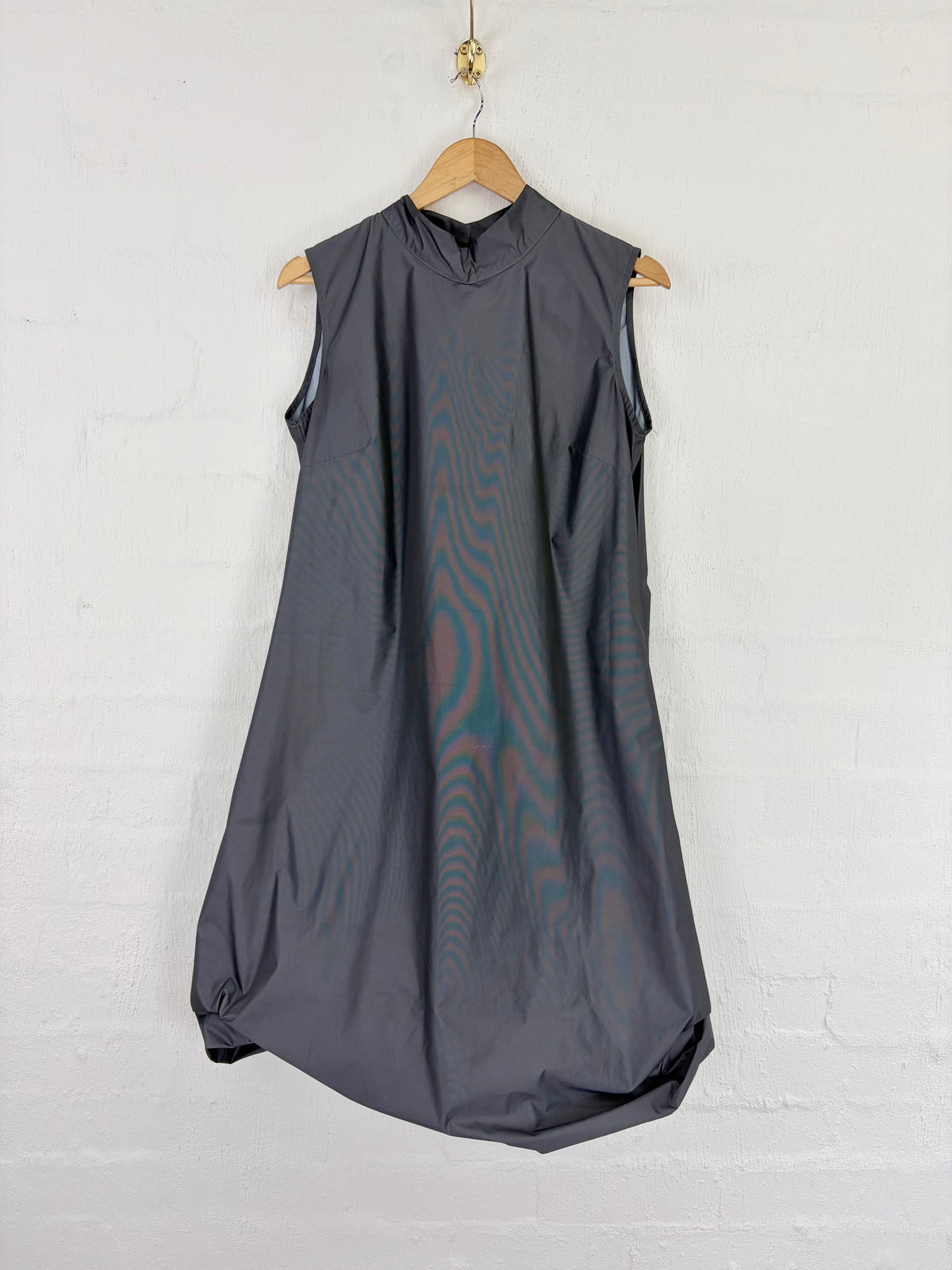 A sleeveless dark gray dress with a high mock neck, hanging on a wooden hanger against a white painted brick wall.