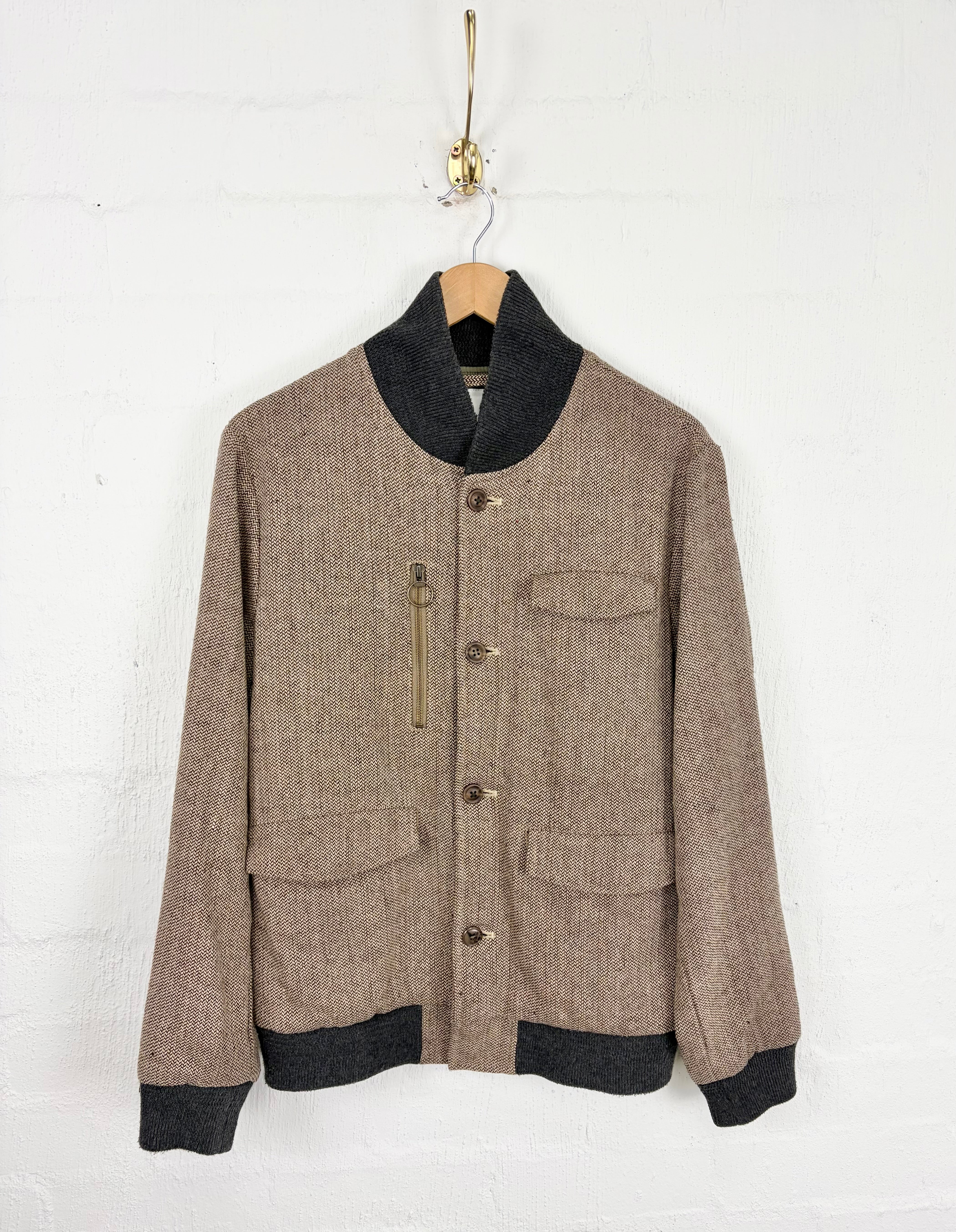 A vintage-style brown tweed jacket with black ribbed knit collar, cuffs, and hem, hanging on a wooden hanger against a white brick wall.