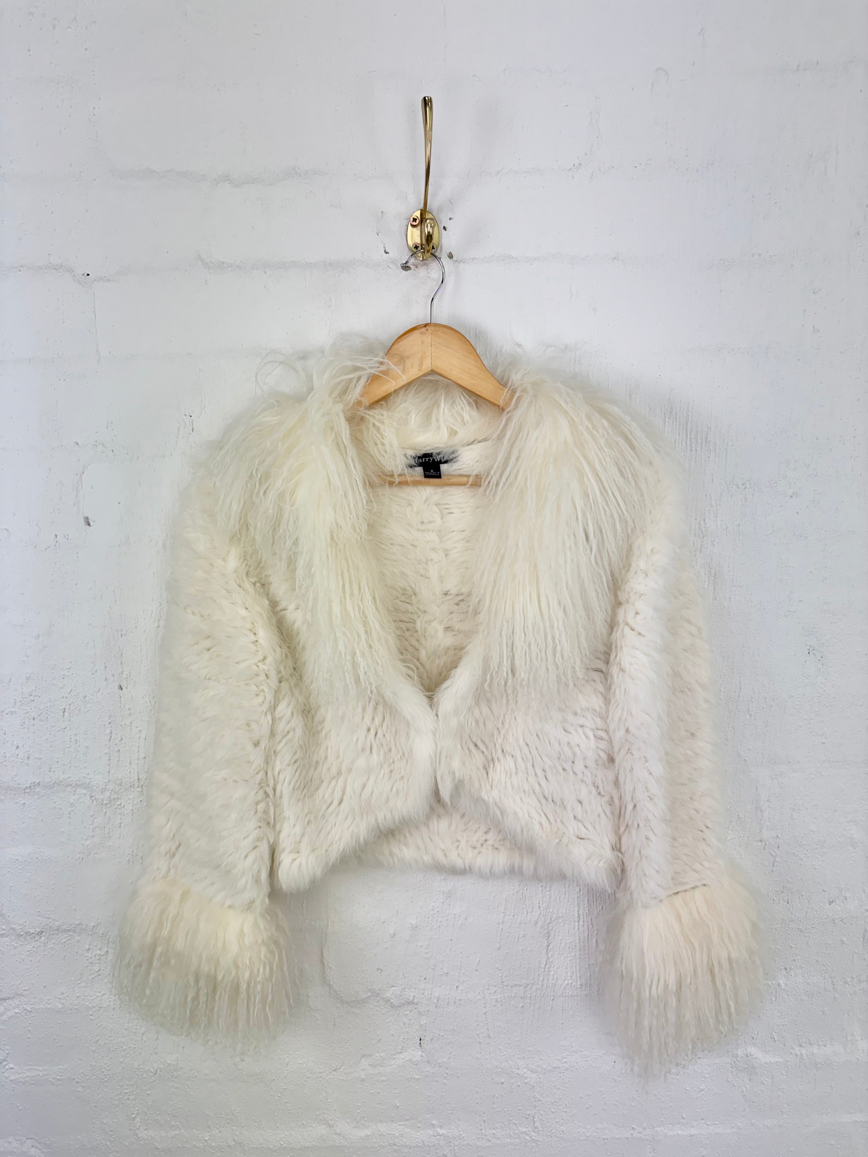 A fluffy off-white faux fur cropped jacket with long sleeves, hanging on a wooden hanger from a gold wall hook against a white brick wall.