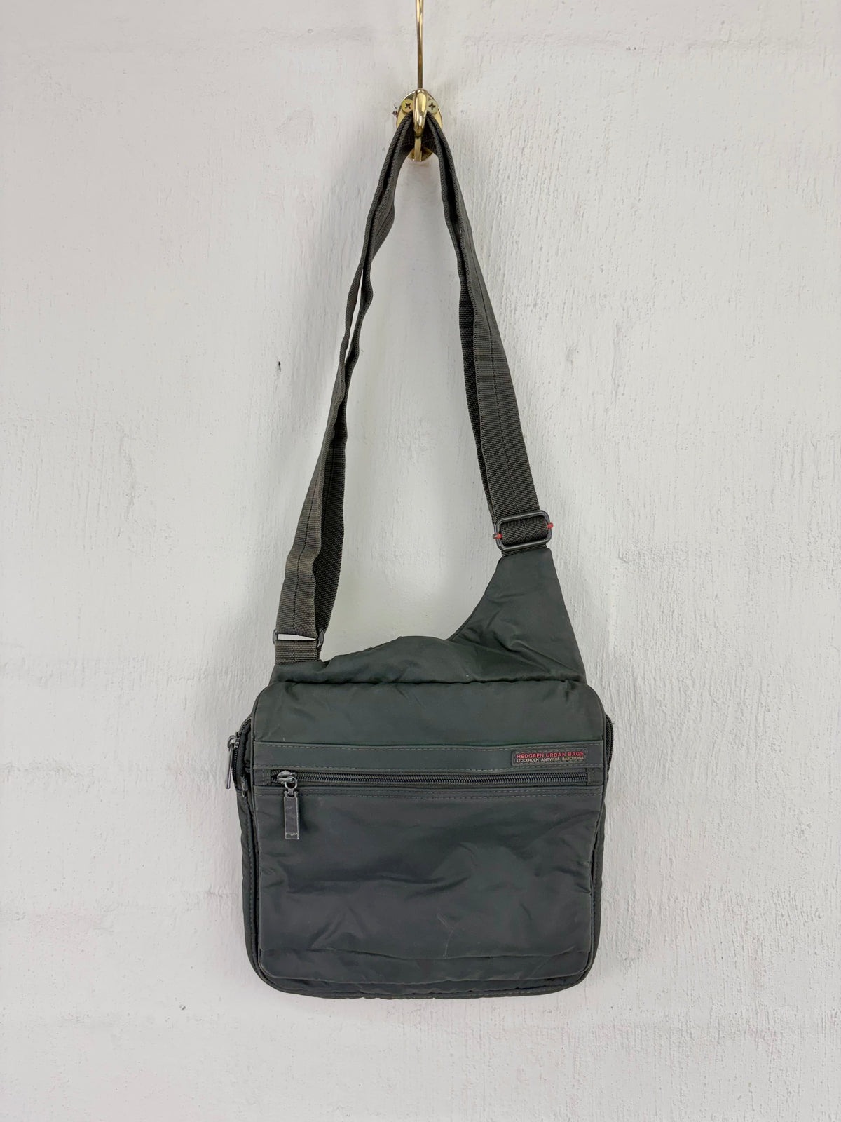 A dark gray nylon shoulder bag with a single adjustable strap, hanging from a gold hook on a white textured wall. The bag features a front zippered pocket with a small red 'BONSAI' brand label.
