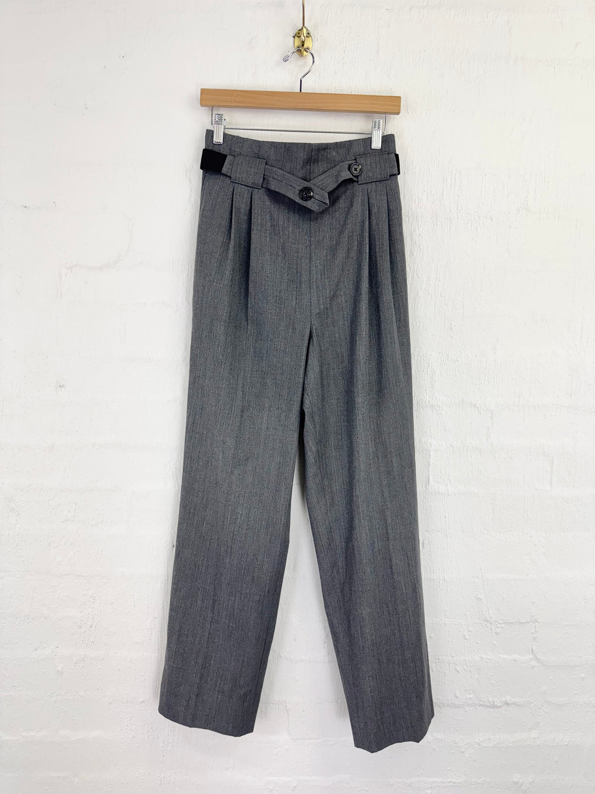 A pair of gray pleated trousers hanging from a wooden hanger against a white painted brick wall.