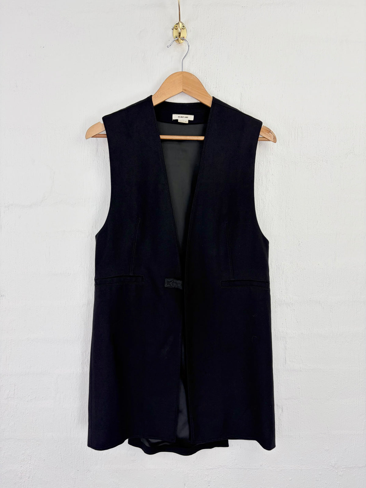 A sleeveless black vest hanging on a wooden hanger against a white painted brick wall.
