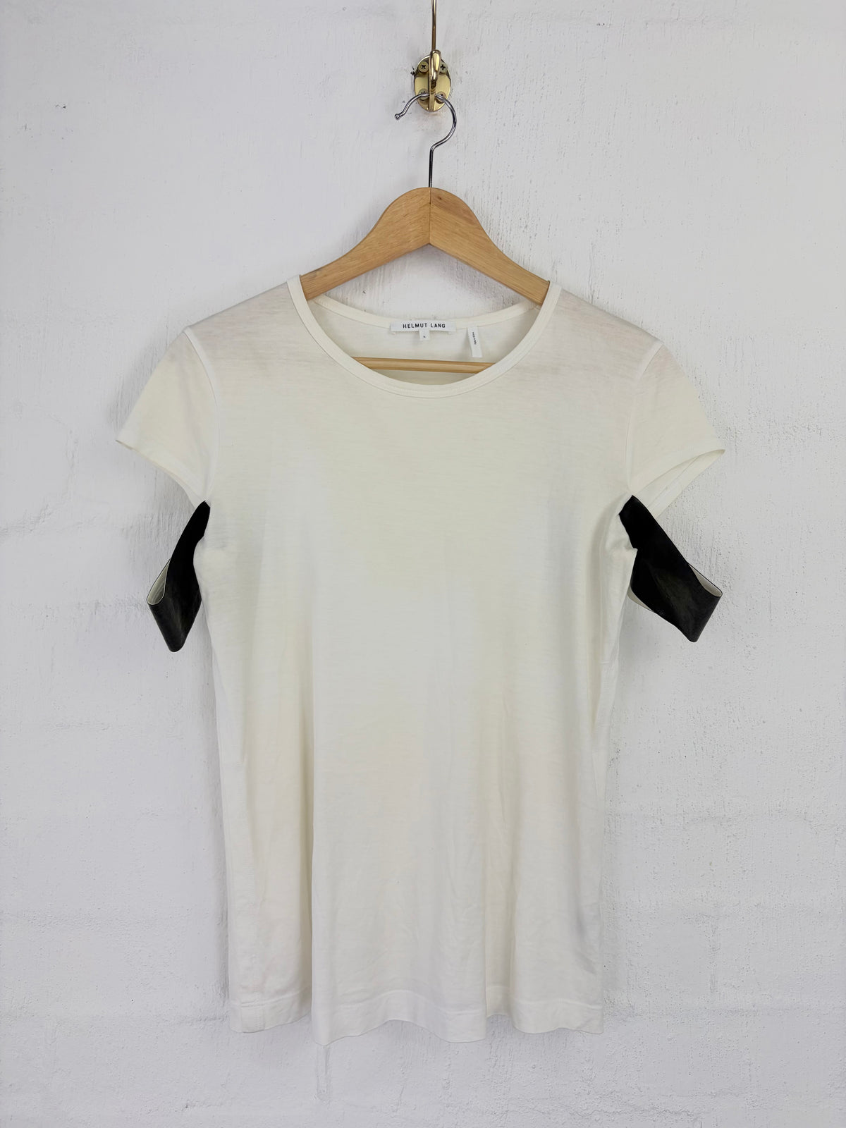 An off-white short-sleeve t-shirt with black faux-leather shoulder accents, hanging on a wooden hanger against a white brick wall.