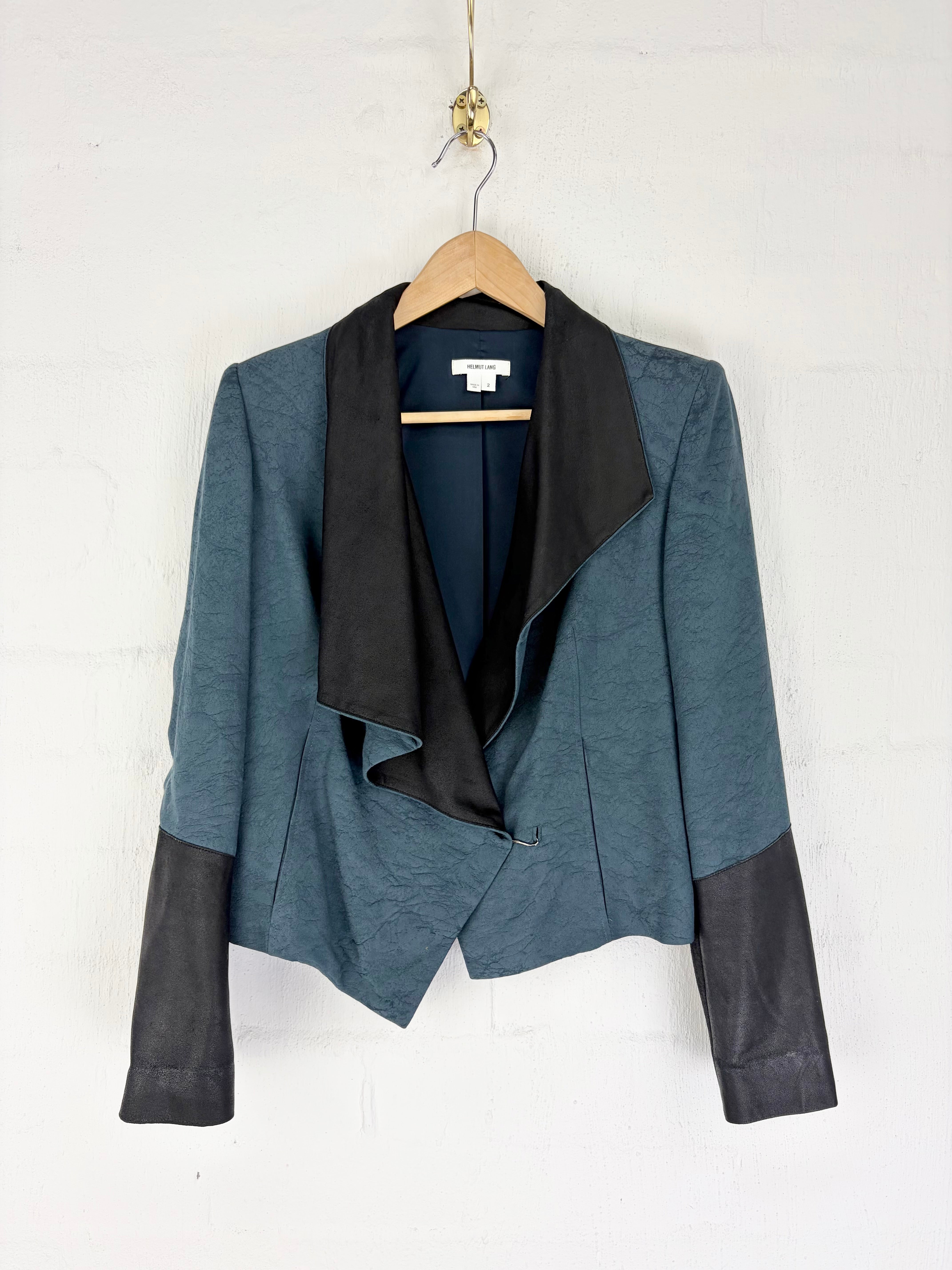 A women's colorblock blazer jacket in slate blue and black, hanging on a wooden hanger against a white painted brick wall.