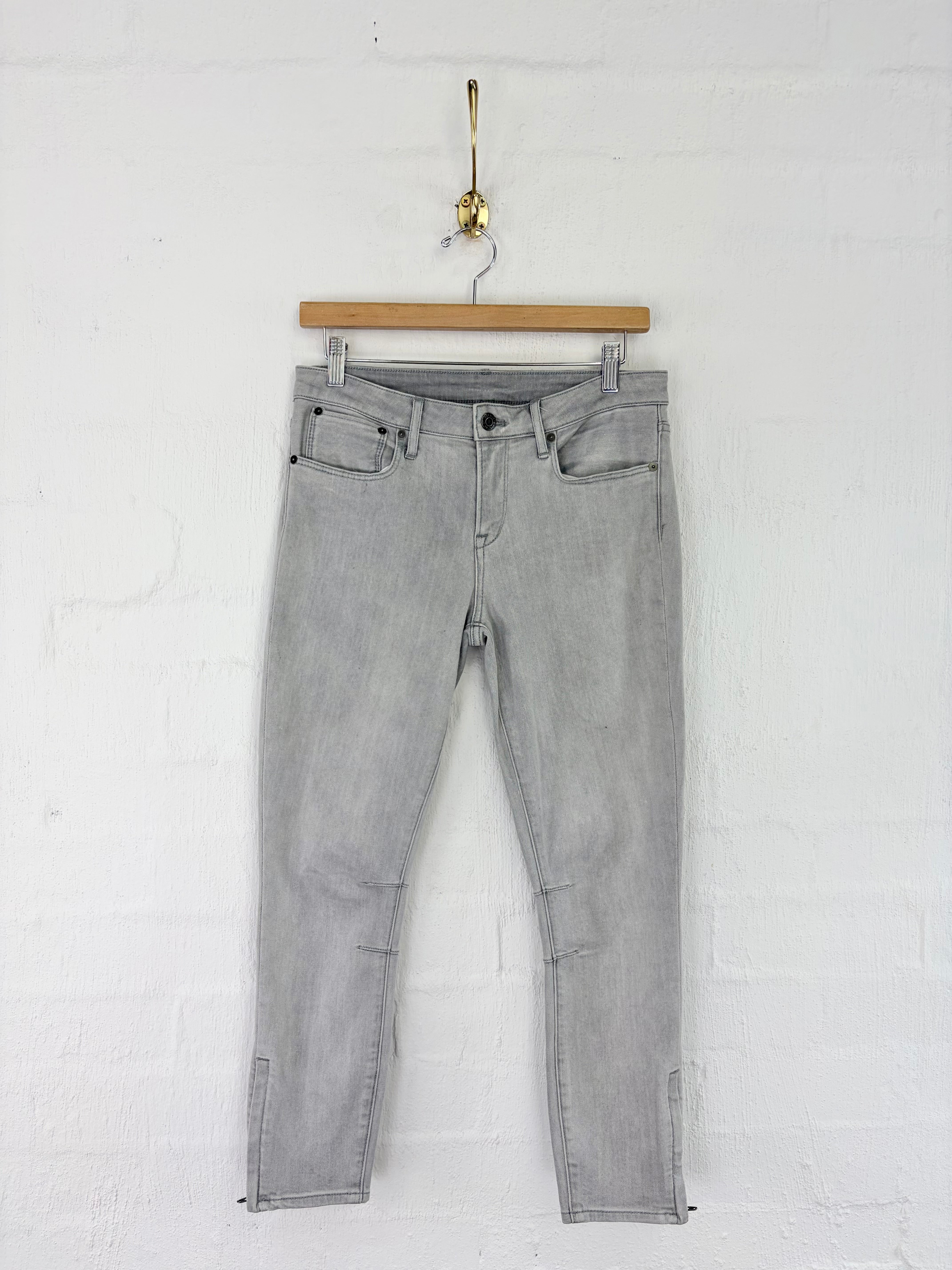 Light gray slim-fit jeans hanging from a wooden hanger on a white brick wall.