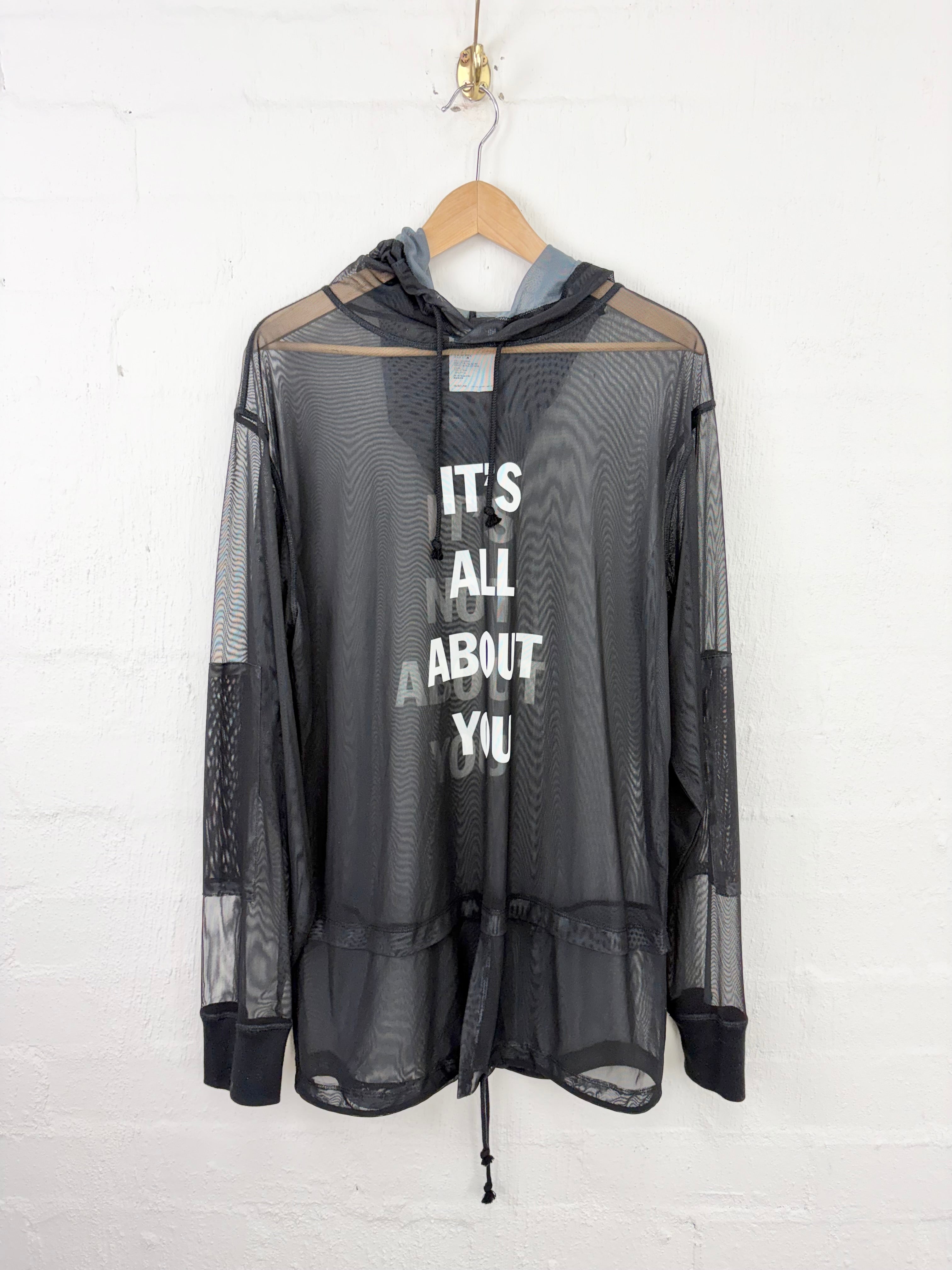 A sheer black mesh hooded long-sleeve top hanging on a wooden hanger against a white painted brick wall. The front has large white text reading 'IT'S ALL ABOUT YOU' and a gray hood lining.