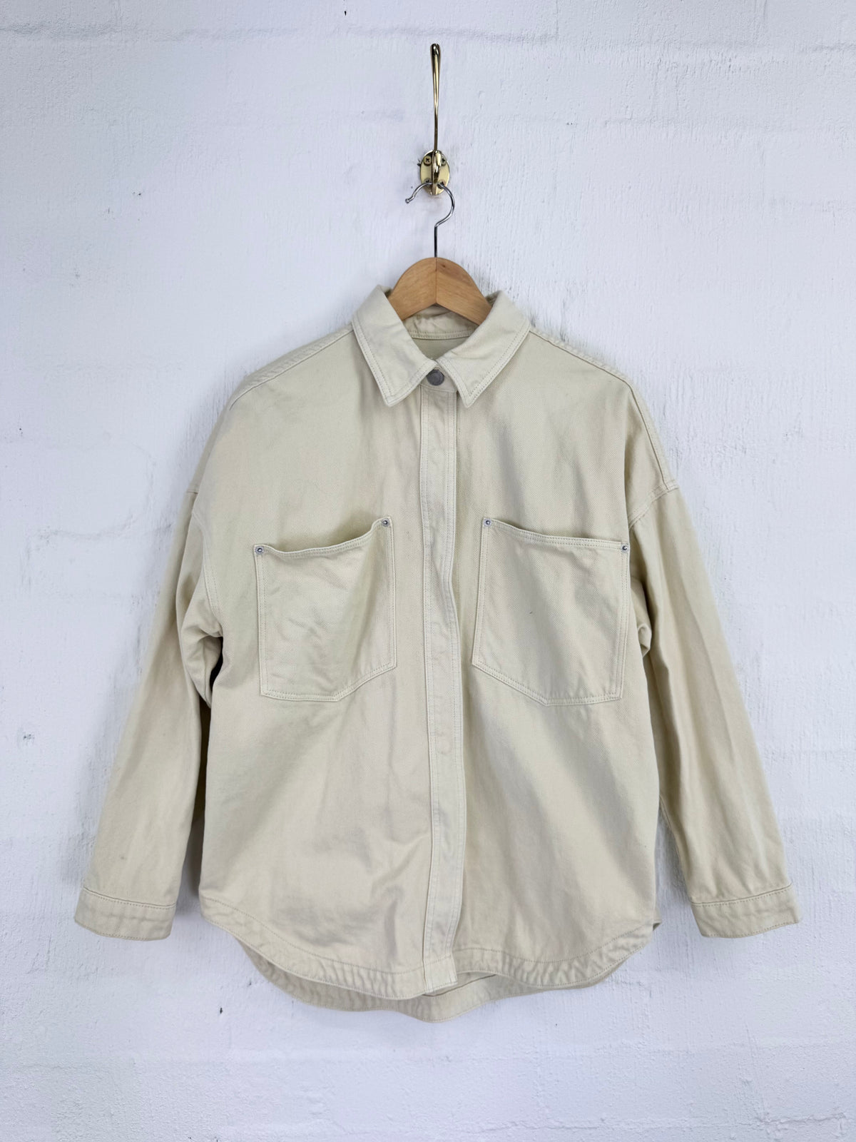 A light beige utility-style button-up shirt with two front chest pockets, hanging on a wooden hanger against a white painted brick wall.