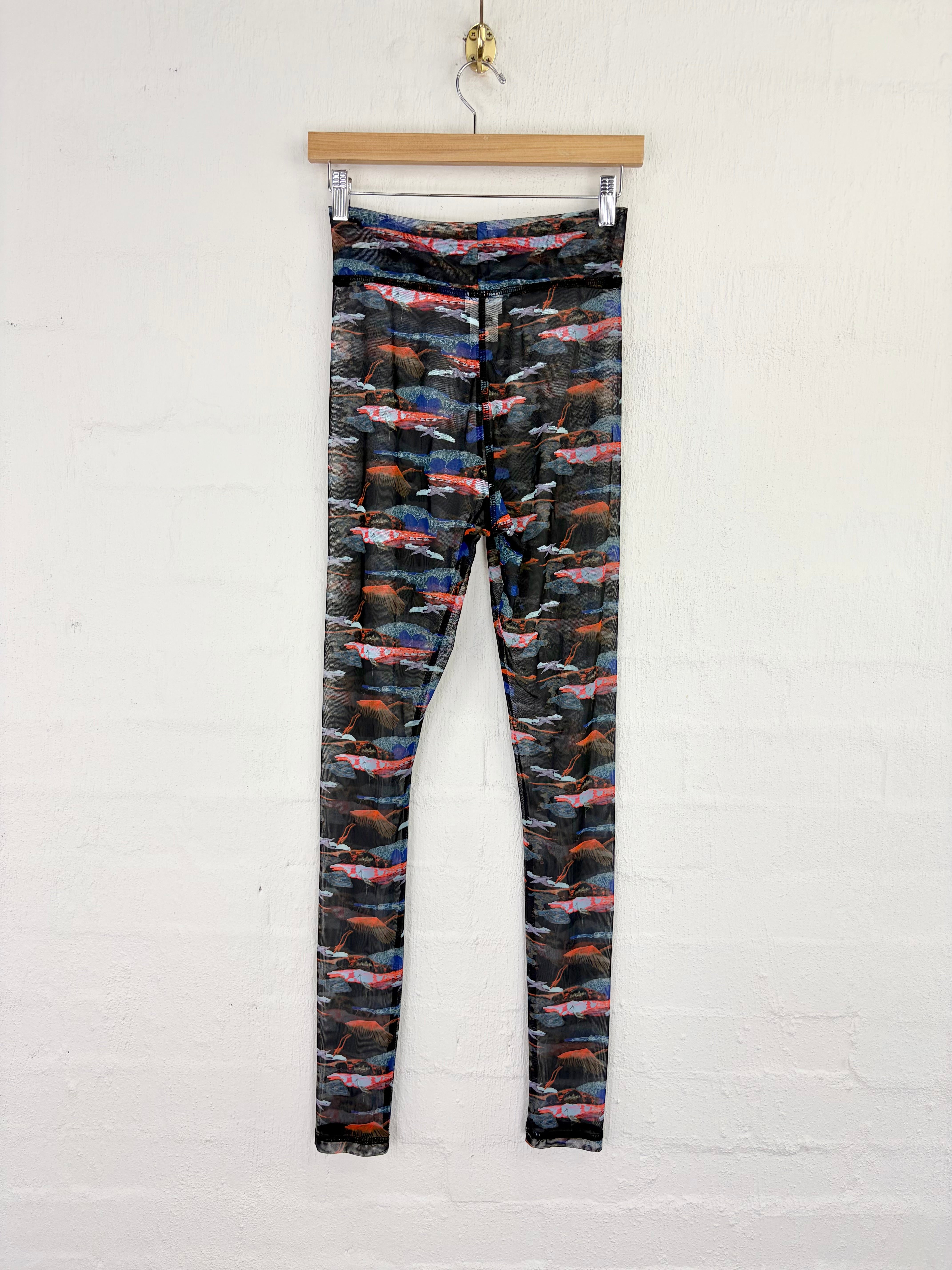 A pair of high-waisted leggings with an abstract multicolor pattern of red, blue, orange, and white brushstrokes on a black background, hanging from a wooden hanger against a white brick wall.