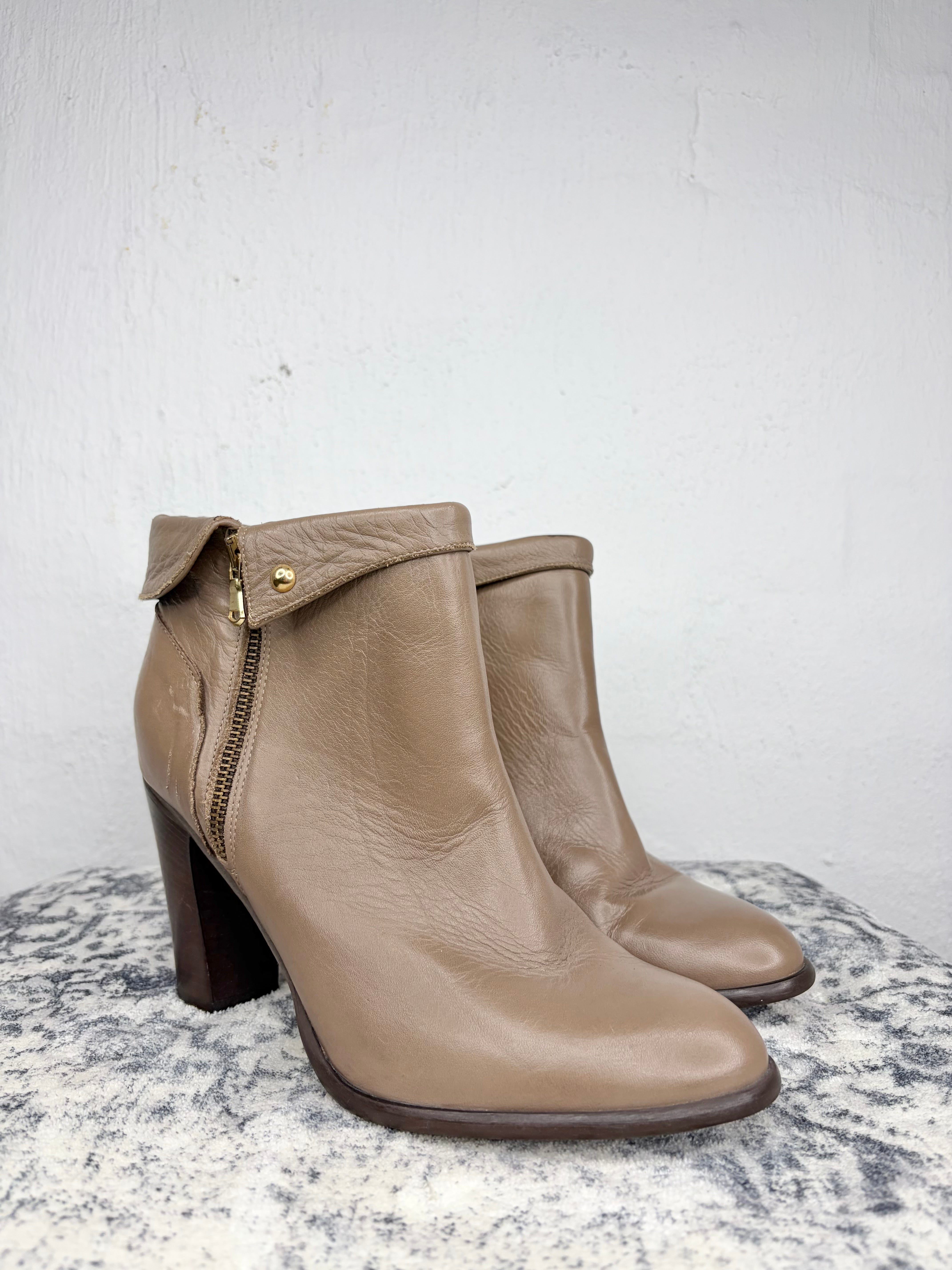 A pair of taupe leather ankle boots with a side zipper and block heel, displayed on a textured gray stone surface against a white wall.