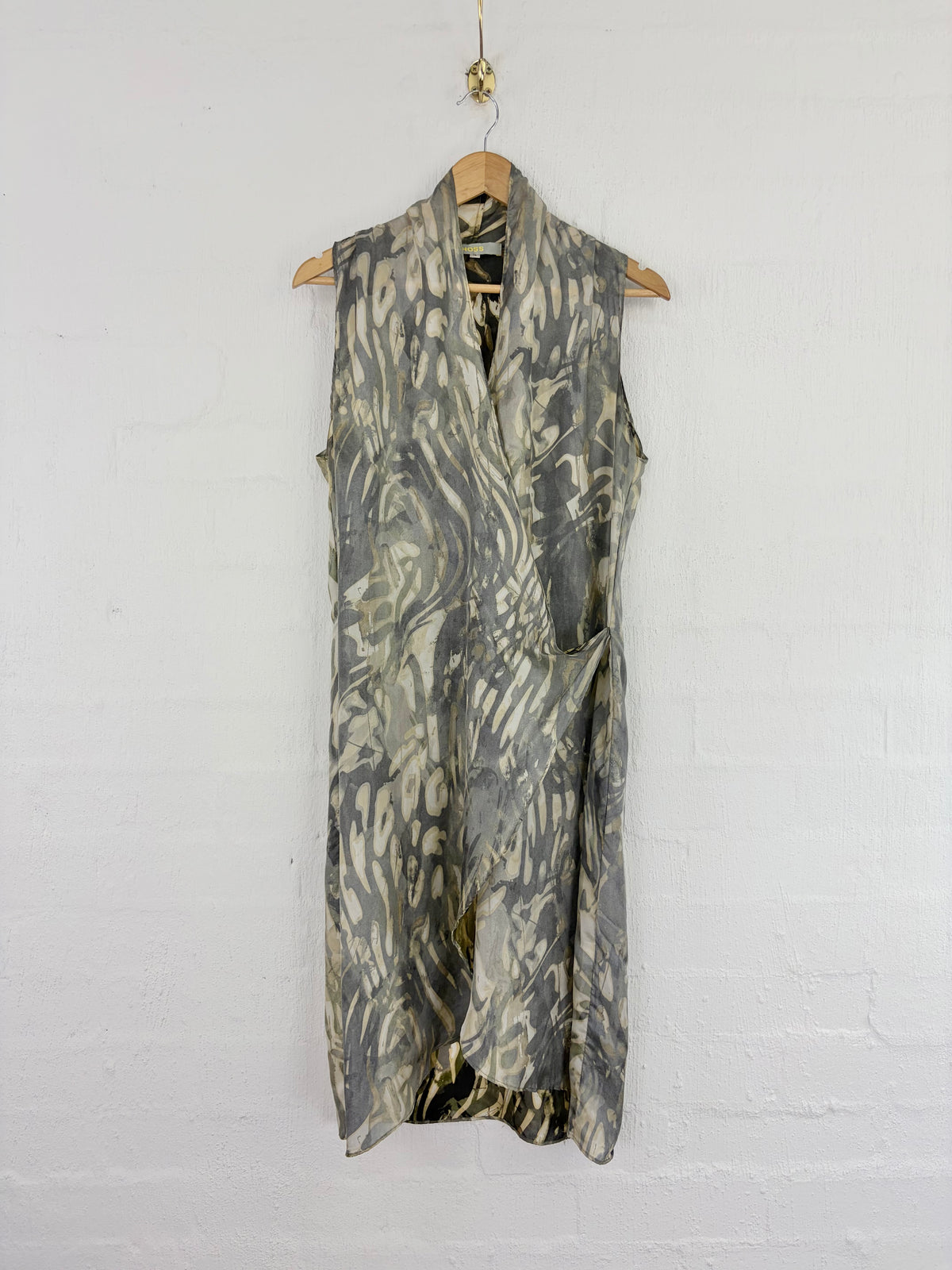 A sleeveless wrap-style dress with an abstract marbled pattern in olive green, gray, and black, hanging on a wooden hanger against a white painted brick wall.