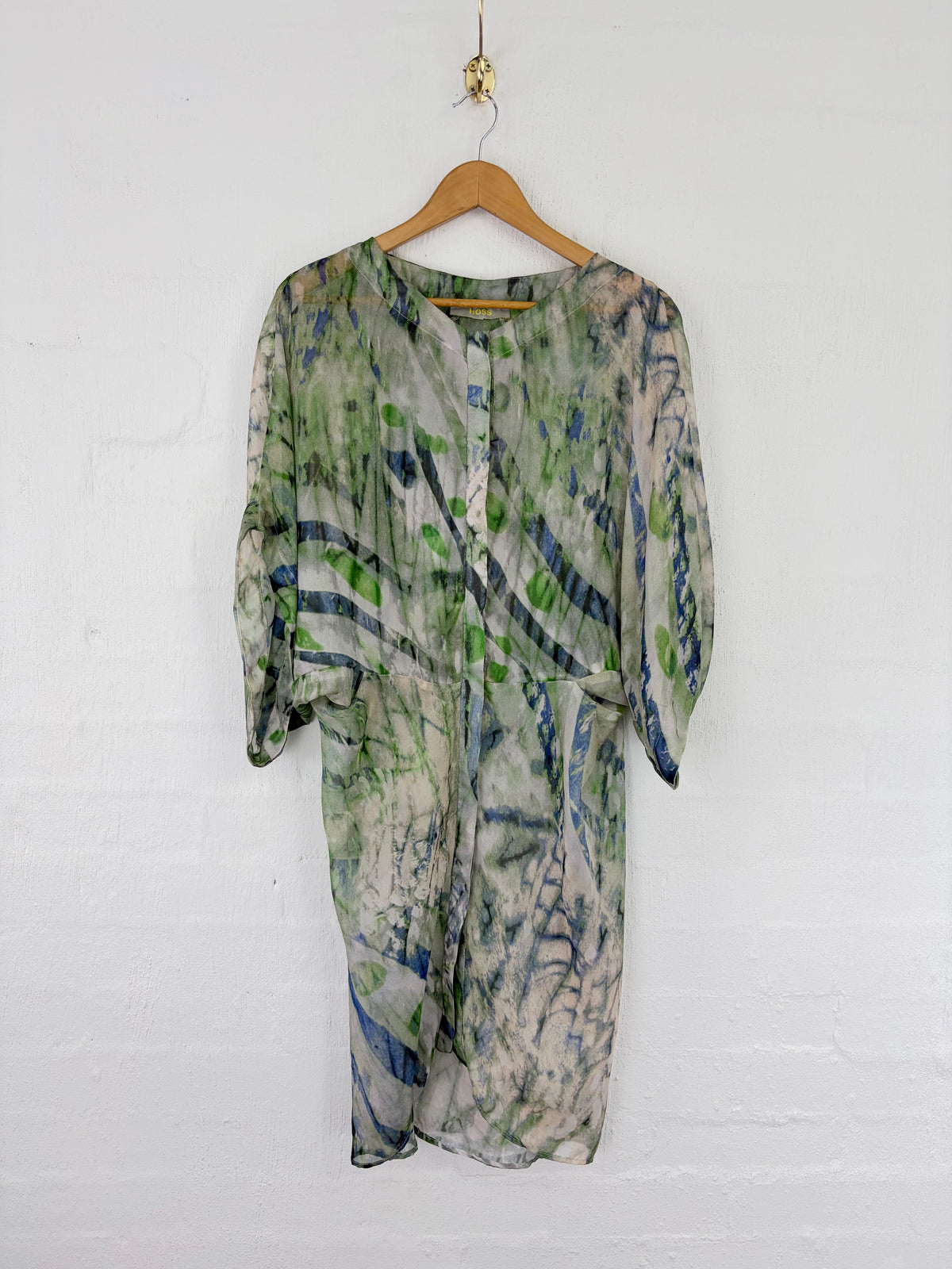 A lightweight, semi-sheer three-quarter sleeve dress with an abstract watercolor print in green, blue, and gray on a cream base, hanging on a wooden hanger against a white painted brick wall.