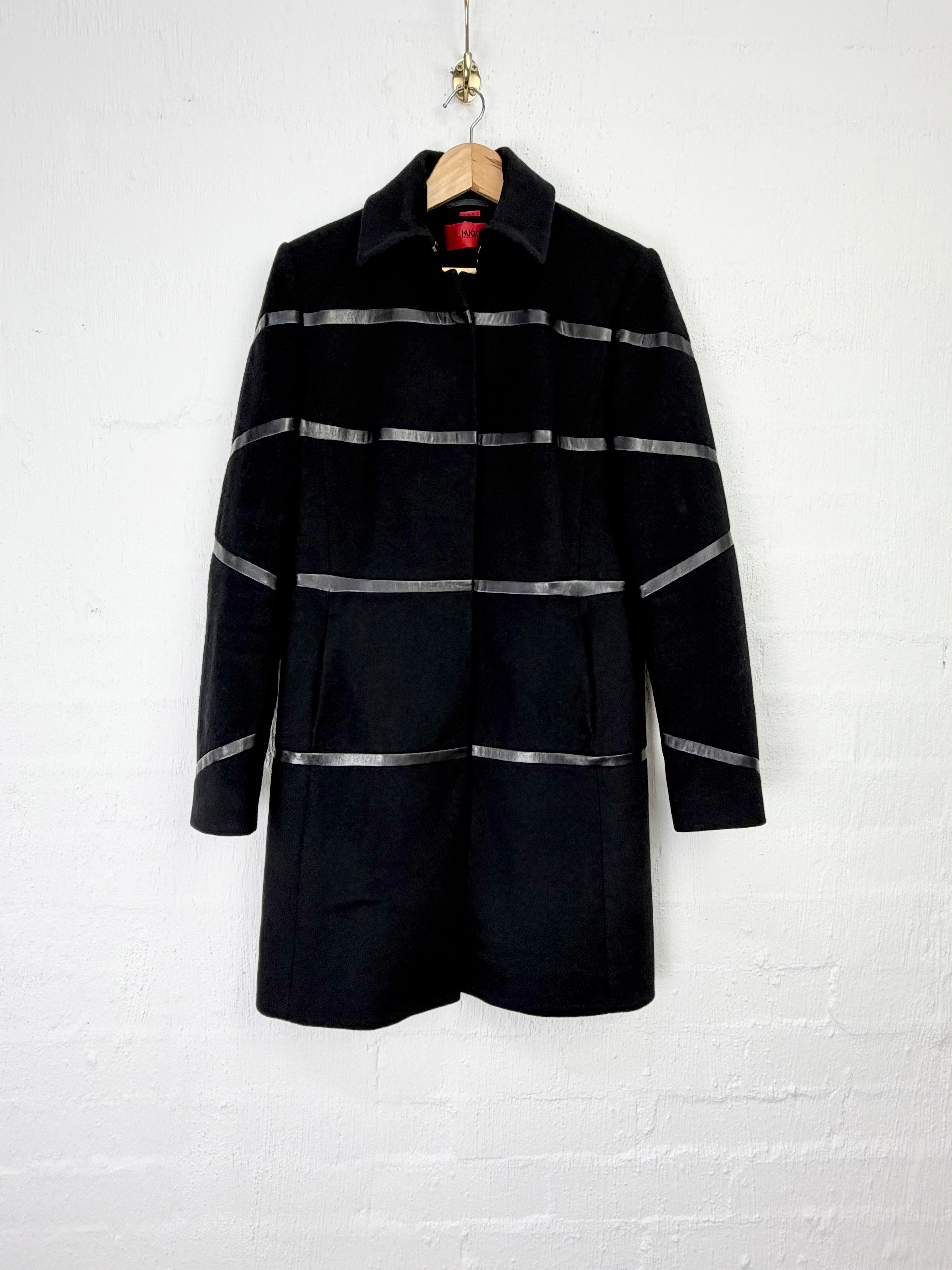 A black wool-blend coat with horizontal metallic silver stripes, hanging on a wooden hanger against a white painted brick wall.