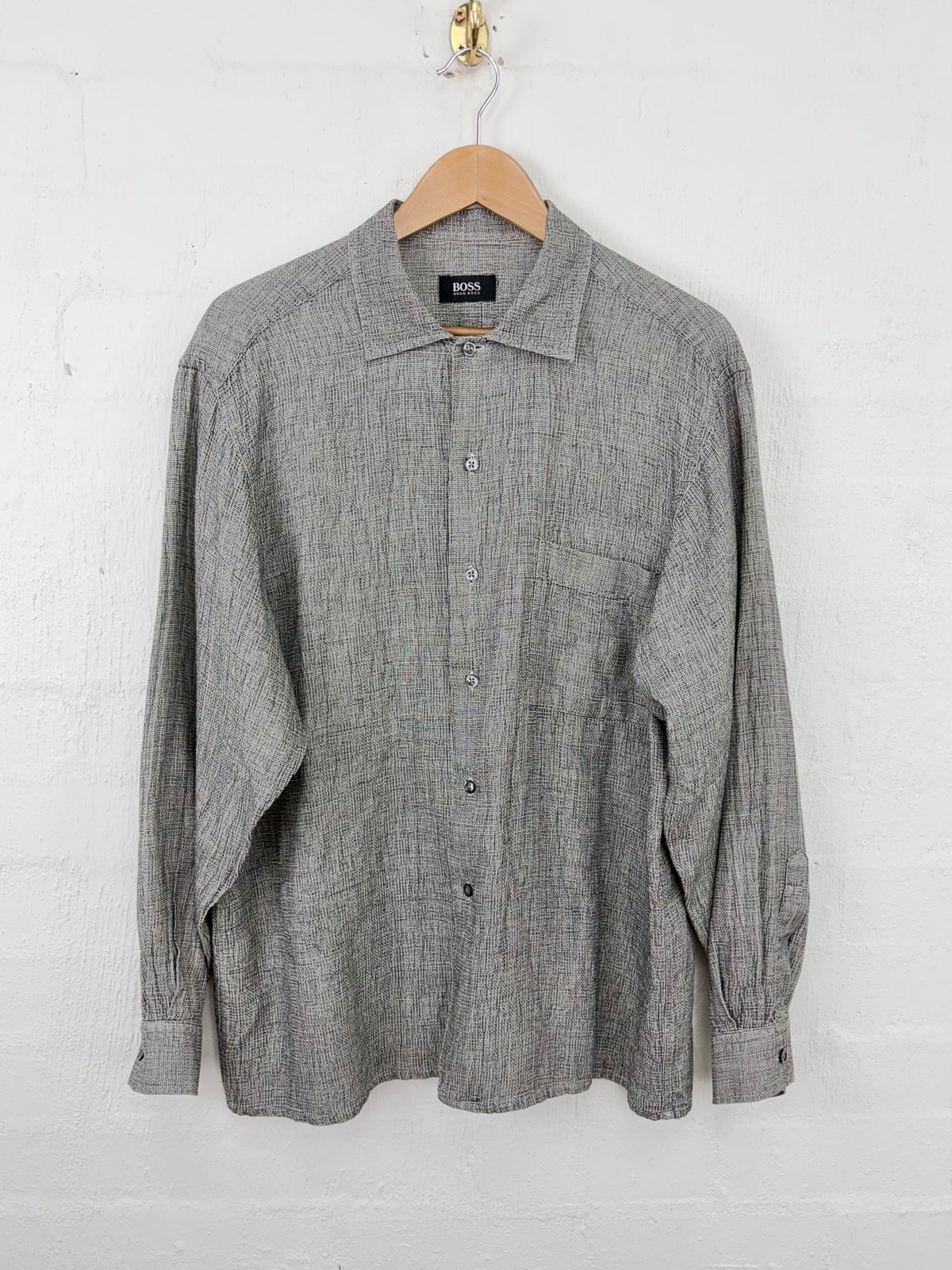 A gray textured long-sleeve button-up shirt with a pointed collar and chest pocket, hanging on a wooden hanger against a white painted brick wall.