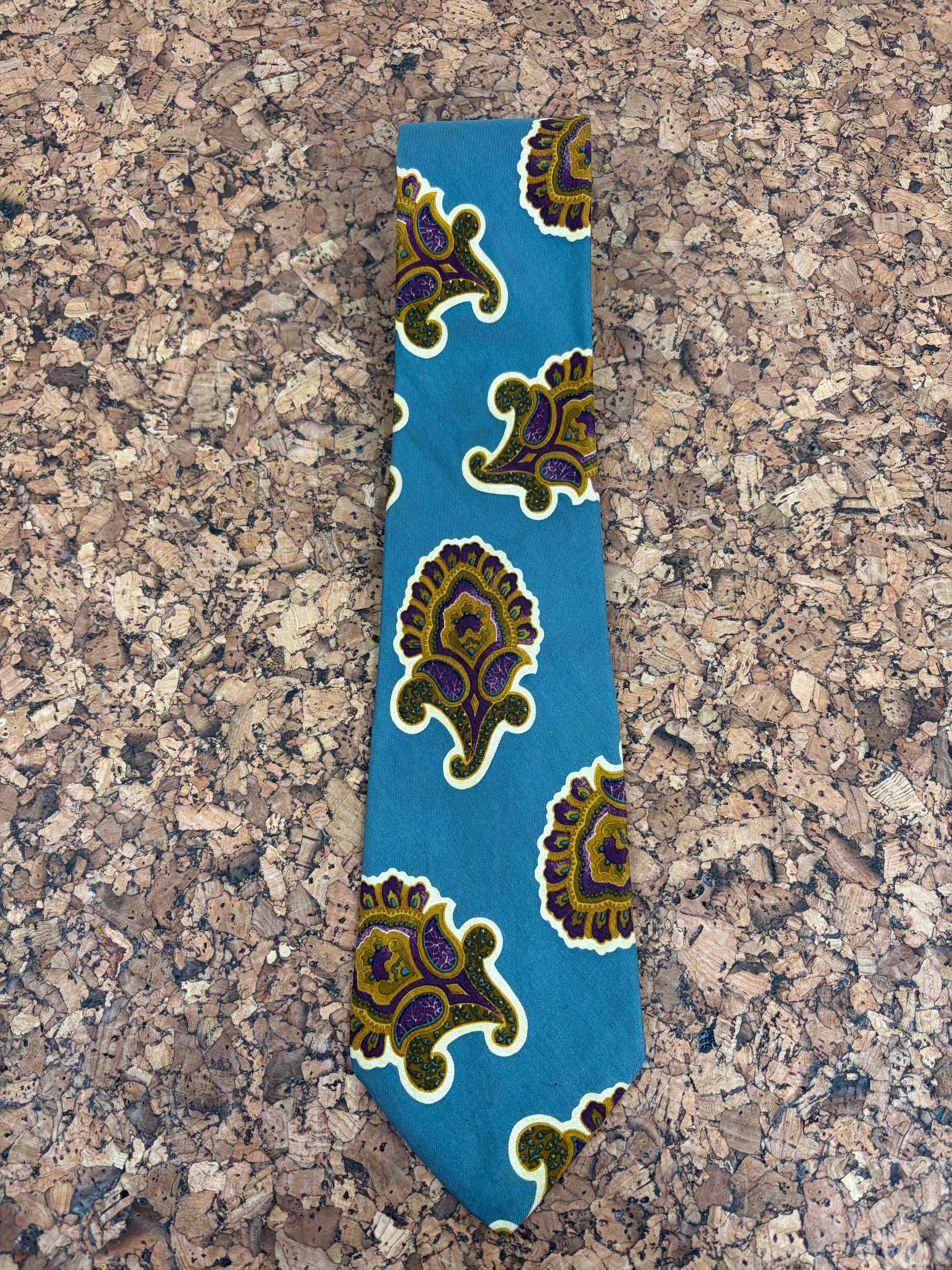 A blue necktie with a repeating paisley pattern in olive green, purple, and white, laid flat on a brown cork surface.