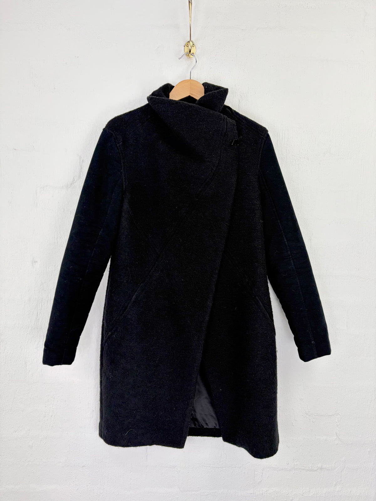 A dark navy blue wrap-style coat hanging on a wooden hanger against a white painted brick wall.