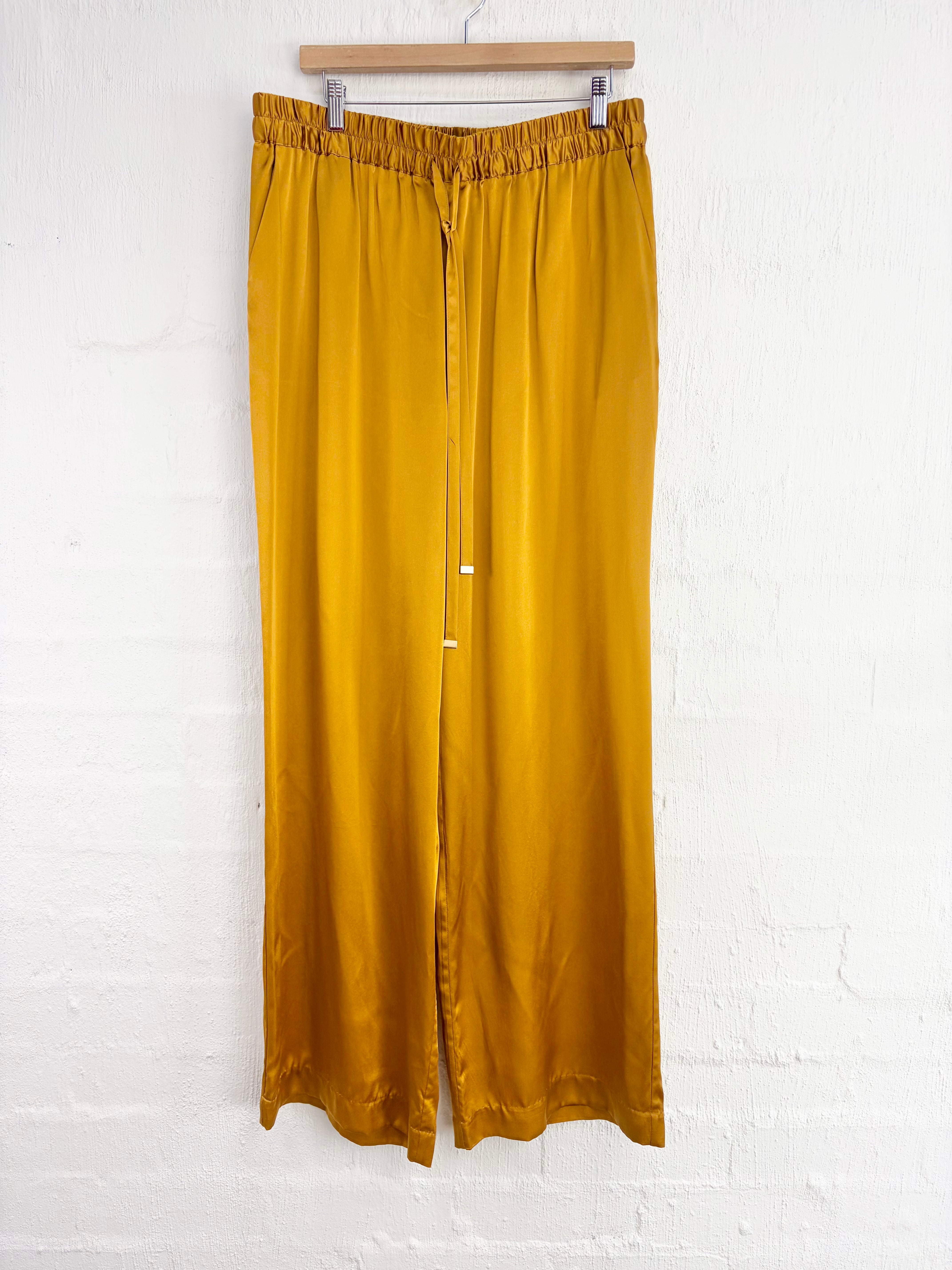 A pair of wide-leg, high-waisted trousers in a rich mustard yellow color, hanging from a wooden hanger against a white painted brick wall.
