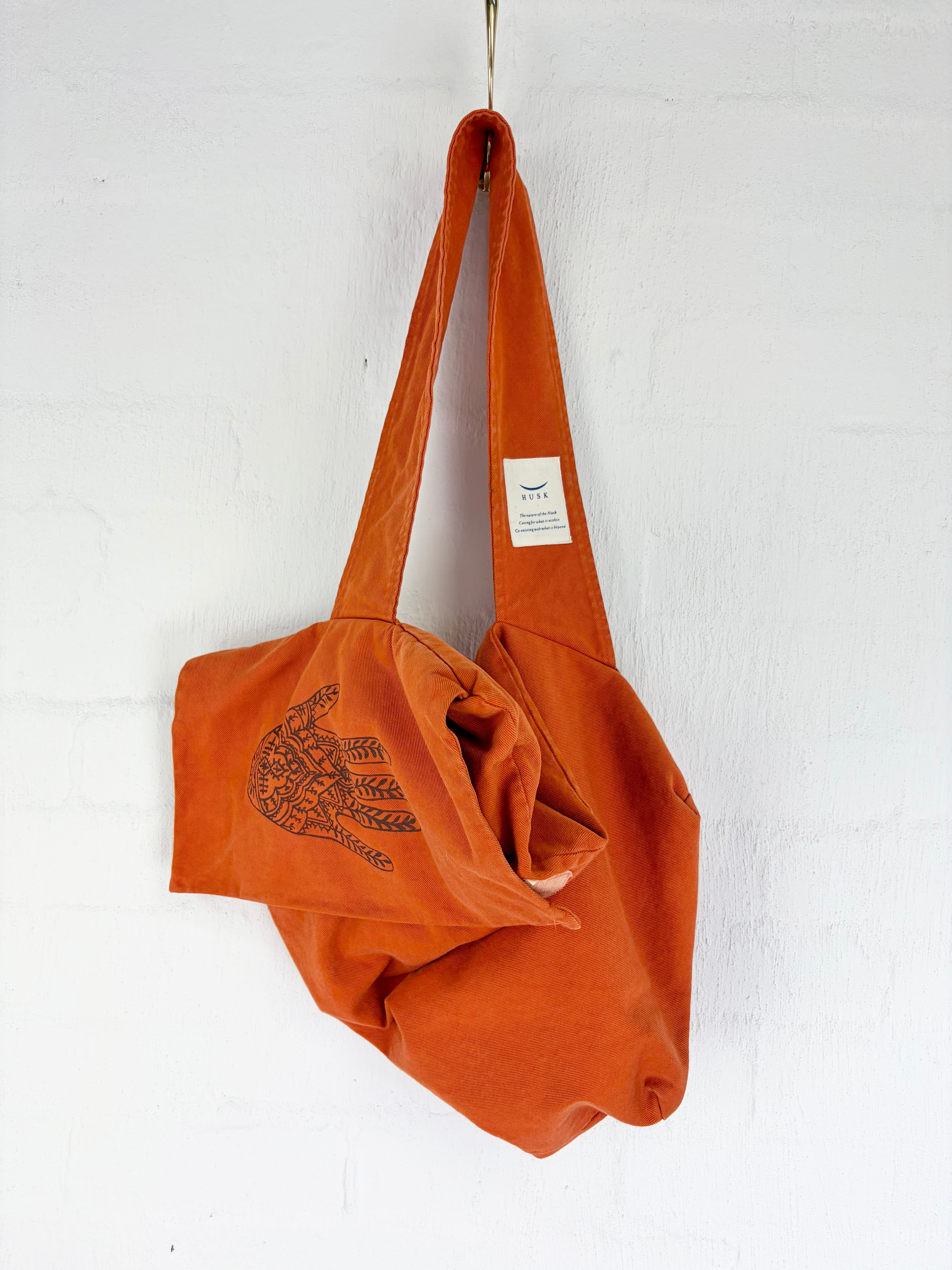 An orange canvas shoulder bag with a black hand-drawn style graphic on the front flap, hanging from a metal hook against a white painted brick wall.