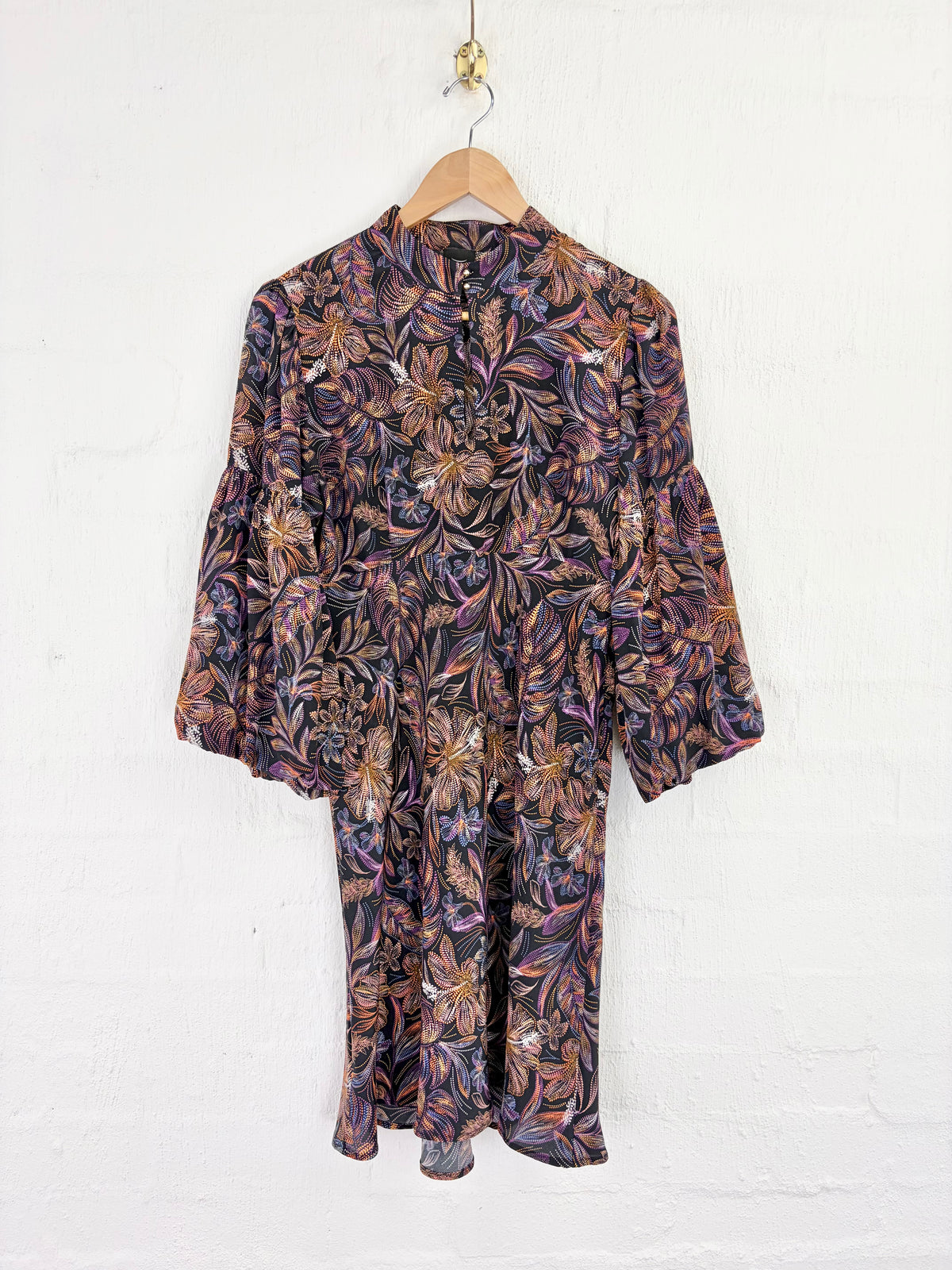 A long-sleeve button-front dress with a dense floral and botanical print in purple, brown, orange, and black, hanging on a wooden hanger against a white painted brick wall.