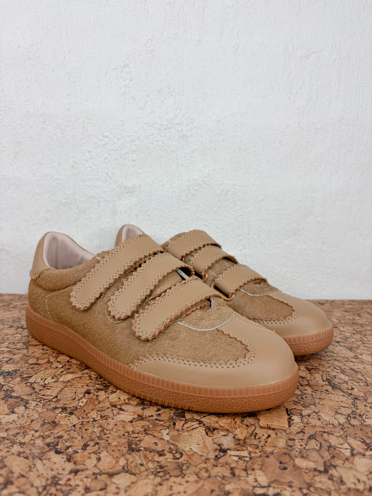 A pair of tan suede and faux-suede low-top sneakers with double hook-and-loop straps, resting on a cork mat against a textured white wall.