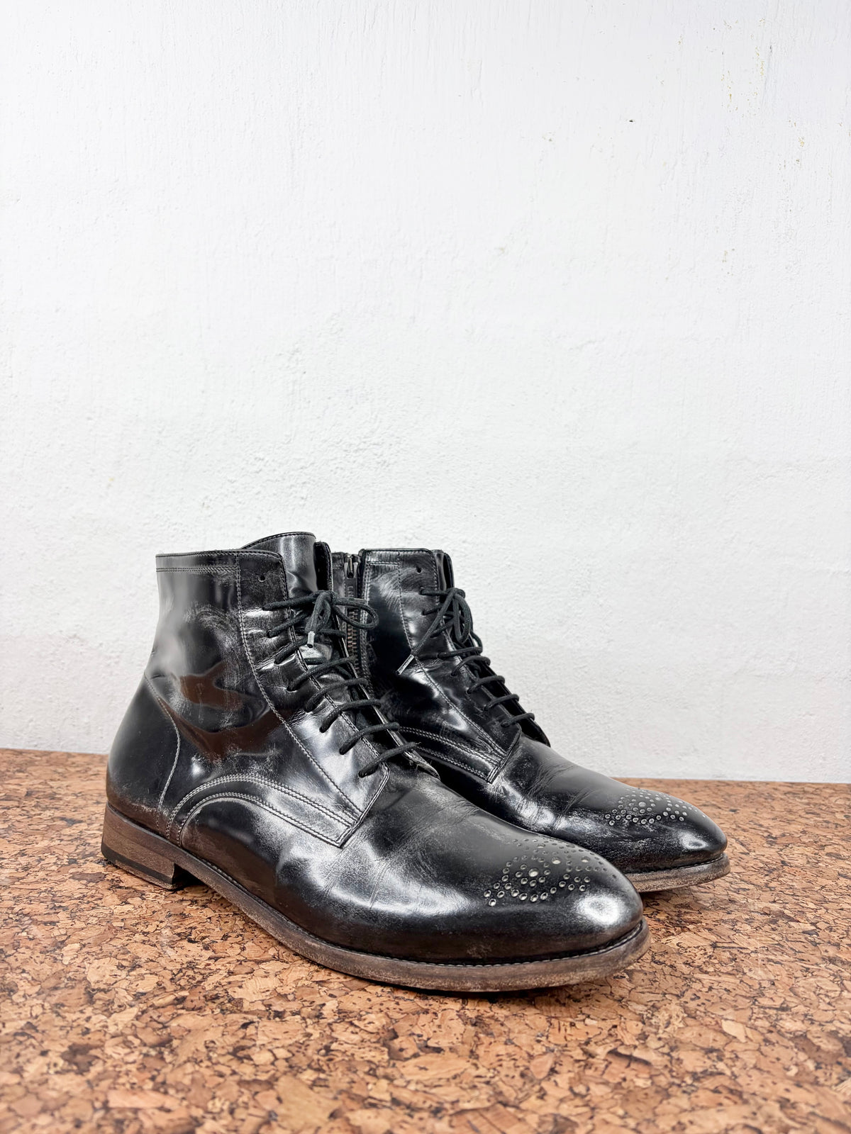 A pair of black leather lace-up ankle boots with brogue detailing on the toes, displayed on a textured brown surface against a white wall.