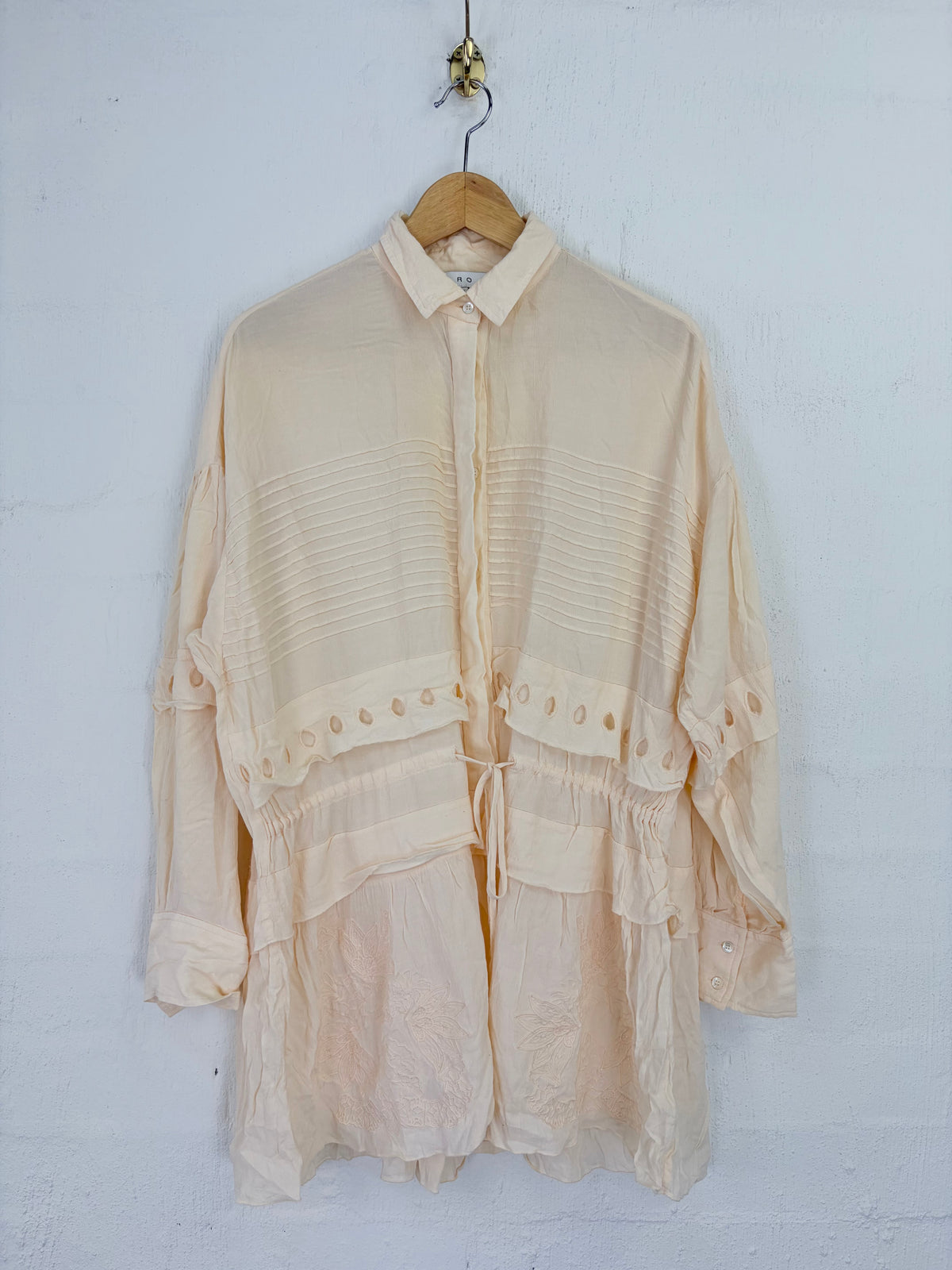 A cream-colored, semi-sheer long-sleeve blouse with pleated details and a tie waist, hanging on a wooden hanger against a white textured wall.