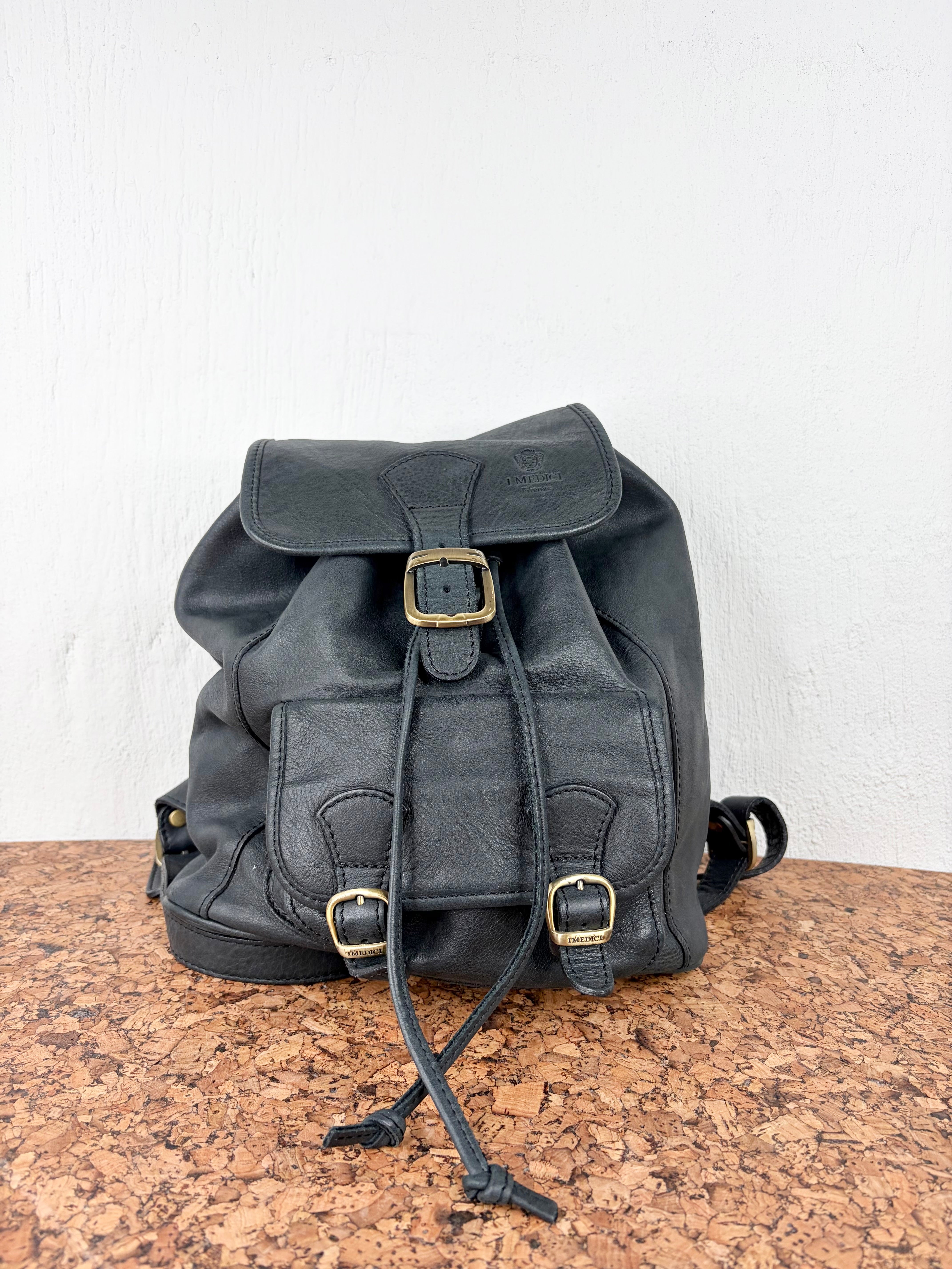 A black leather drawstring backpack with brass-tone buckle details, displayed on a cork surface against a white wall.