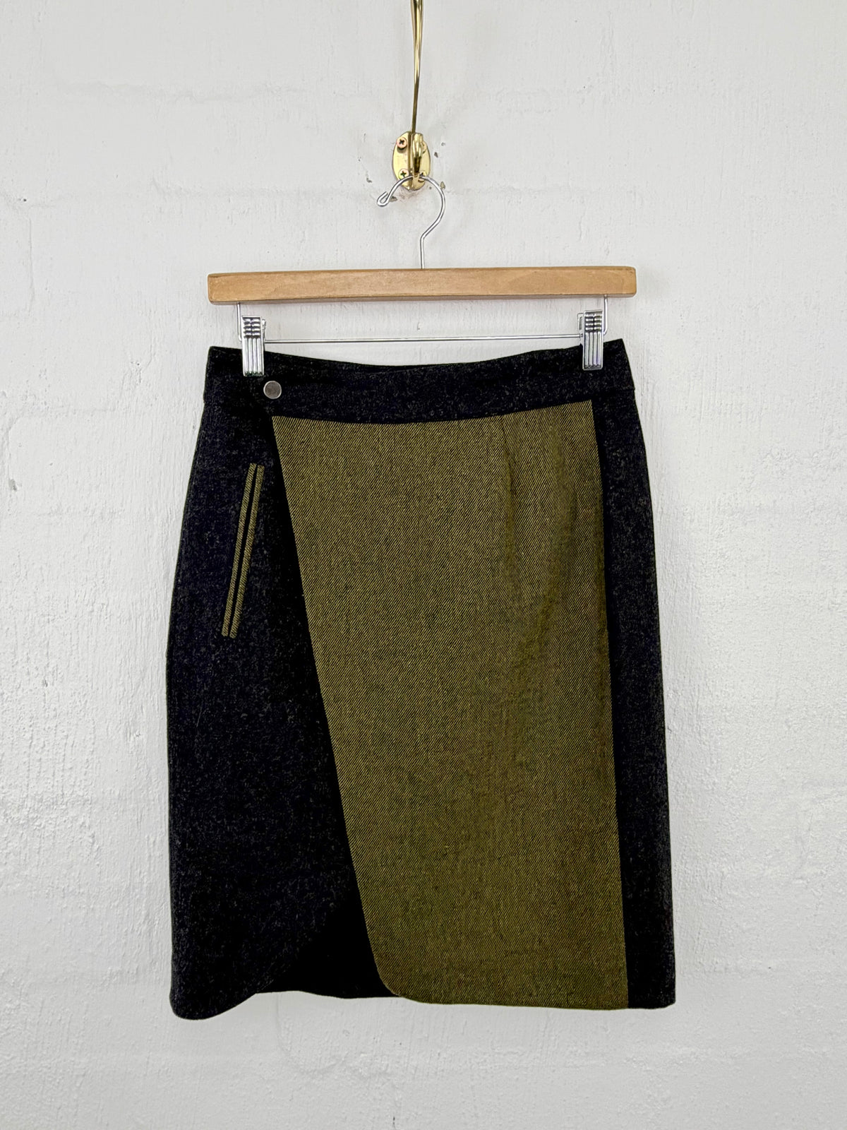 A color-block knit mini skirt in black and olive green, hanging from a wooden hanger against a white painted brick wall.
