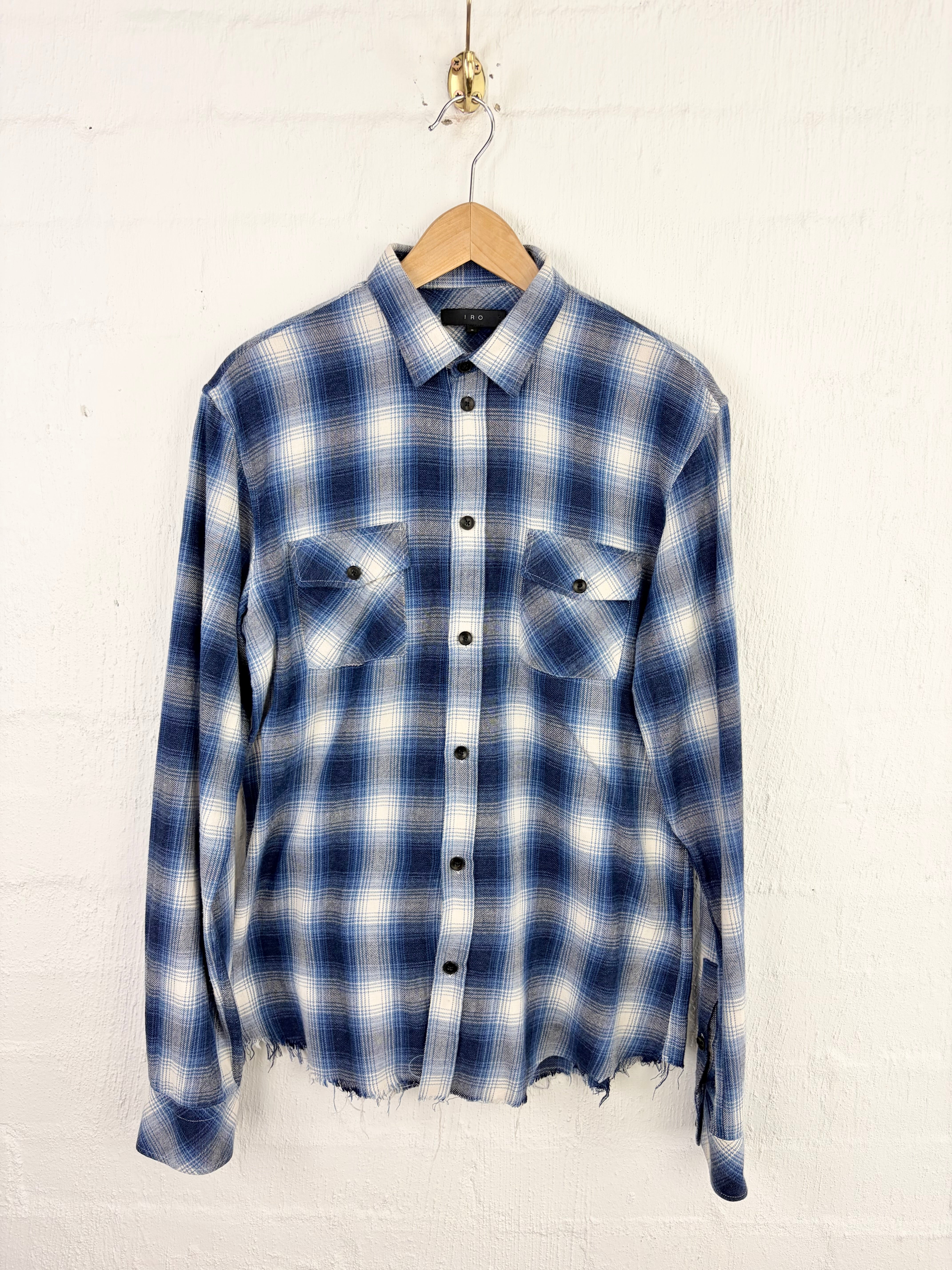 A long-sleeve plaid button-up shirt in blue and white, hanging on a wooden hanger against a white painted brick wall.