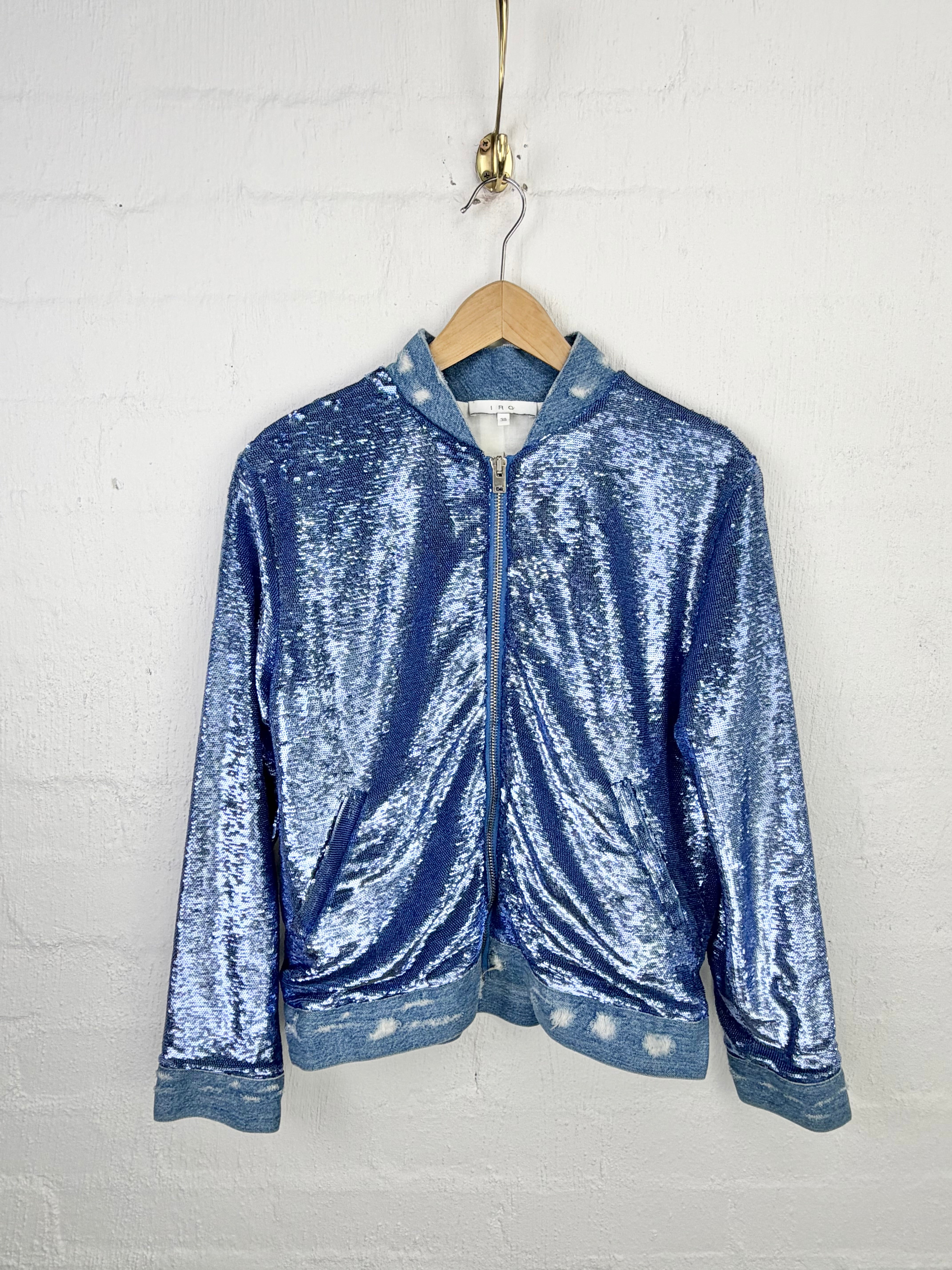 A blue sequined zip-up jacket with a stand collar, hanging on a wooden hanger against a white painted brick wall.