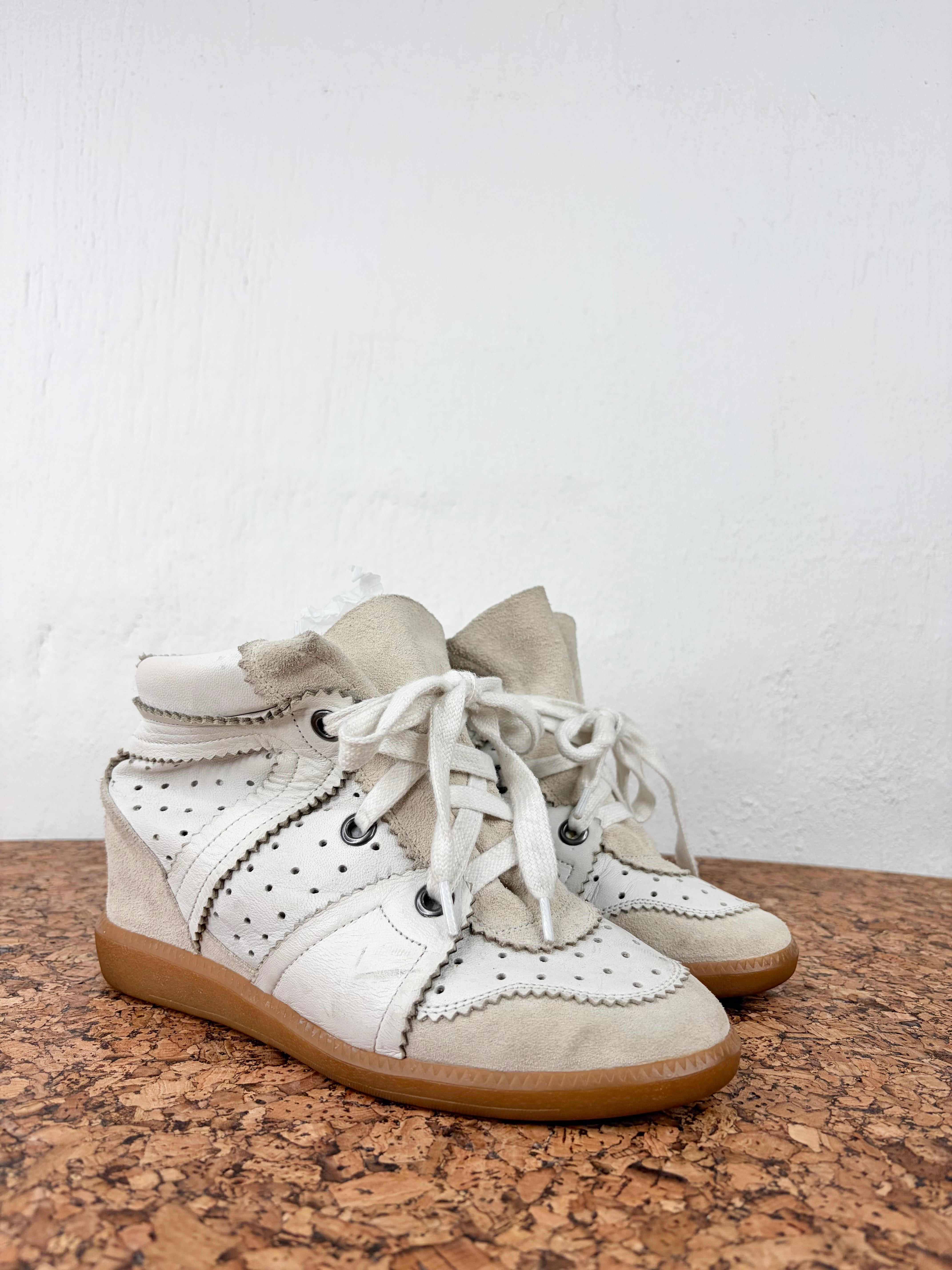 A pair of off-white high-top sneakers with perforated leather panels, white laces, and tan soles, displayed on a cork surface against a white wall.