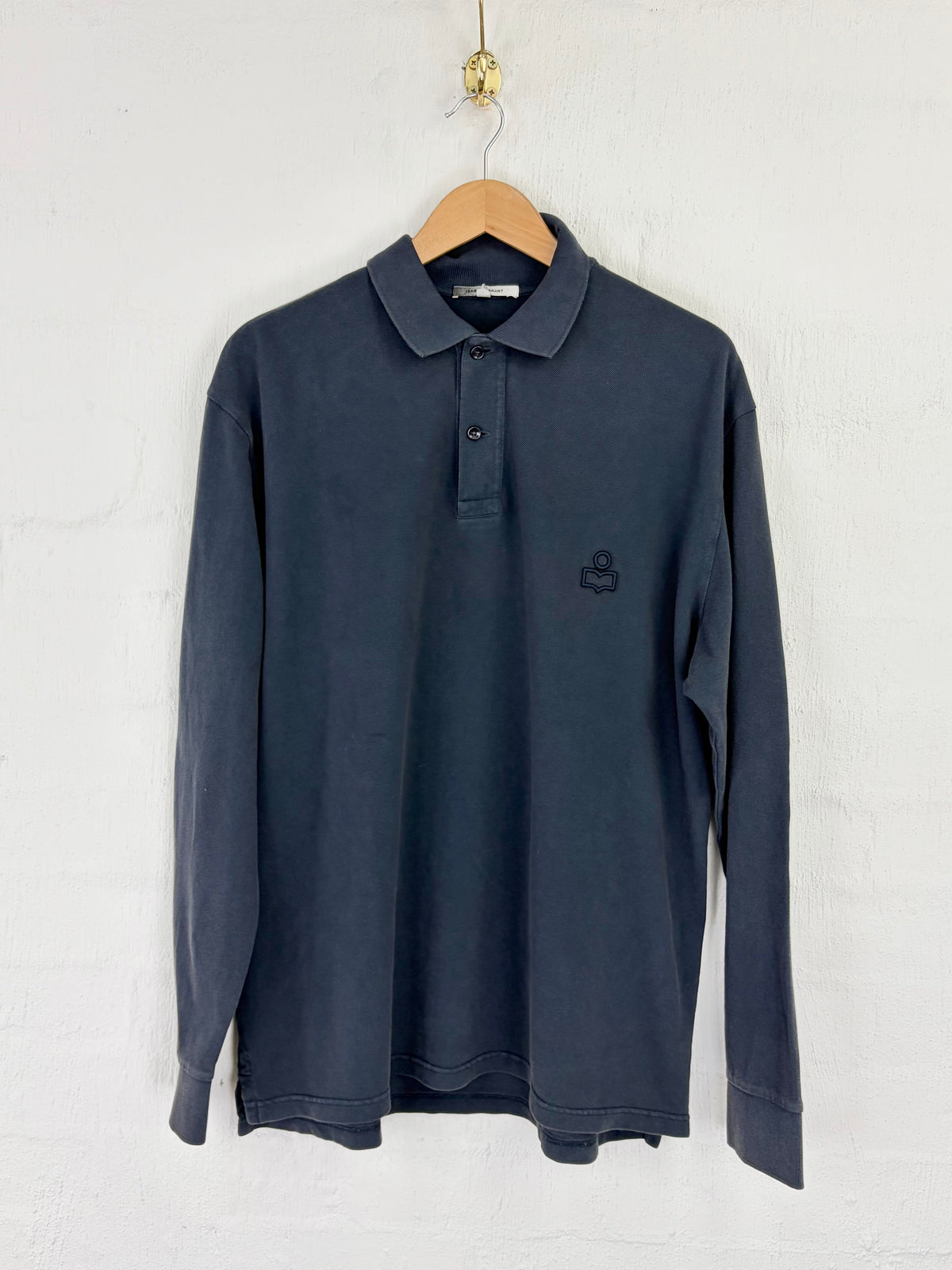 A dark slate blue long-sleeve polo shirt hanging on a wooden hanger against a white textured wall. The shirt features a small dark embroidered logo on the left chest.