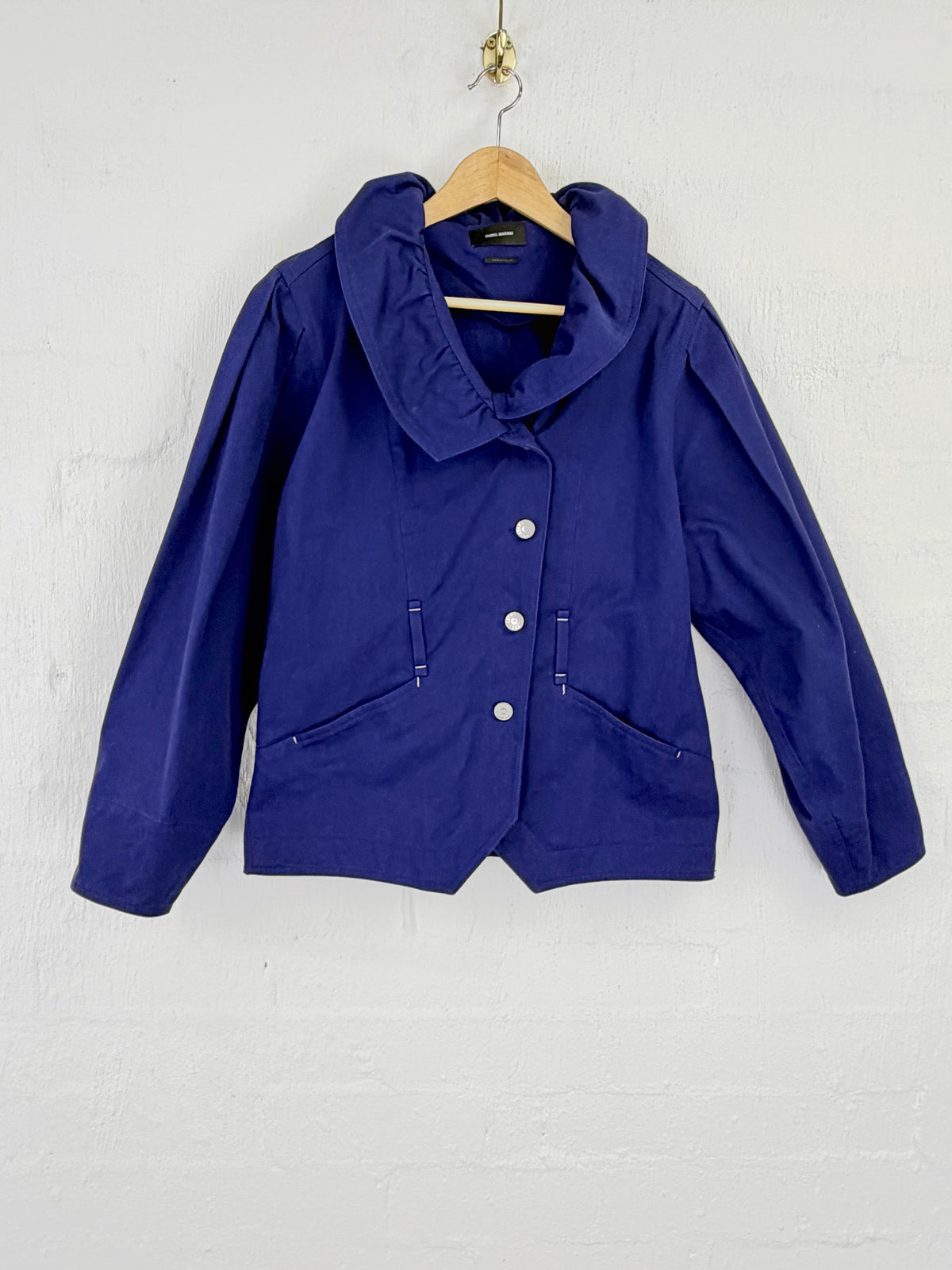 A royal blue cropped jacket with a wide collar and three front buttons, hanging on a wooden hanger against a white painted brick wall.