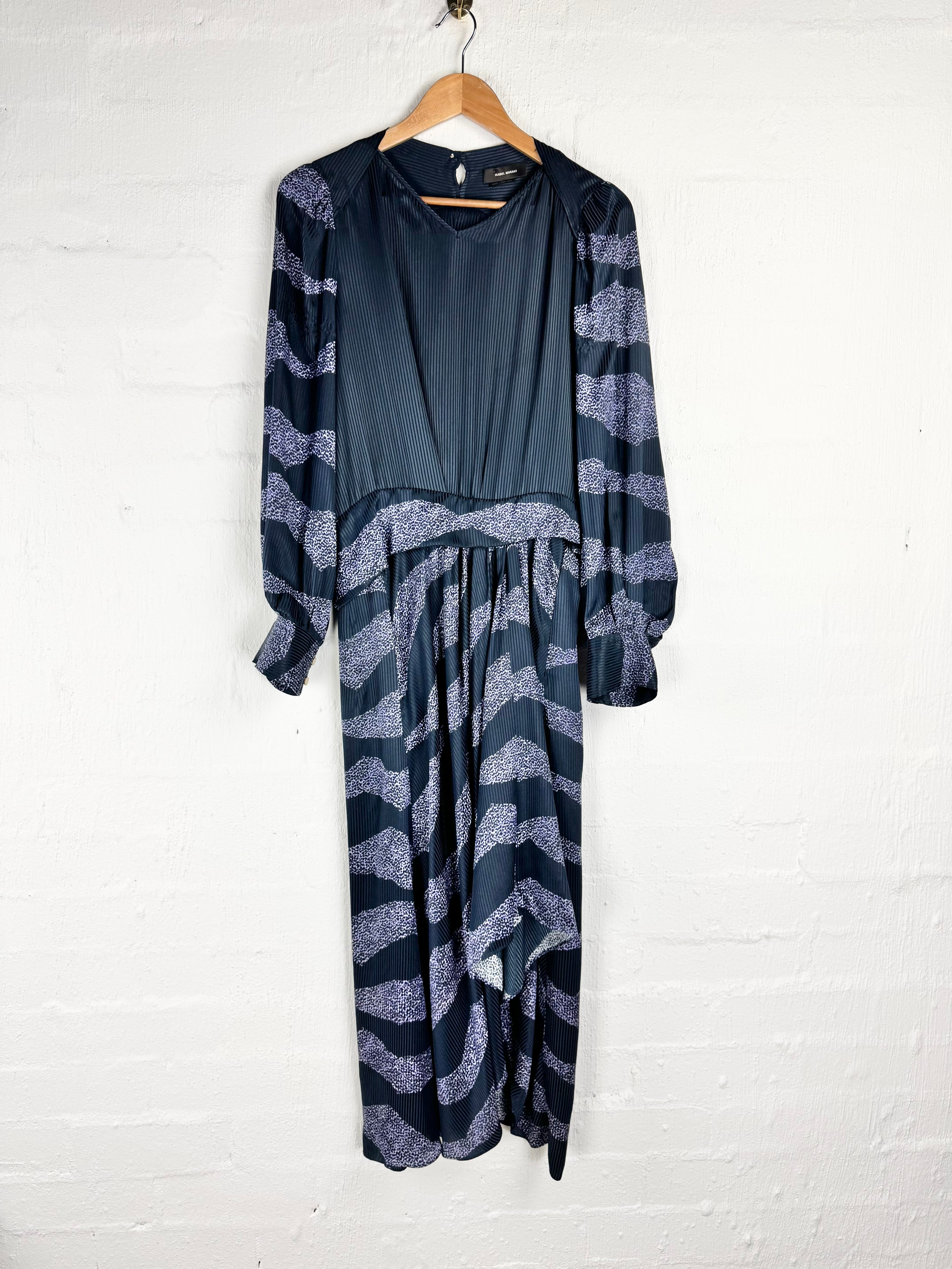 A long-sleeved maxi dress with a solid dark navy blue bodice and a flowing skirt featuring a wavy, abstract pattern in navy and light lavender-gray, hanging on a wooden hanger against a white painted brick wall.