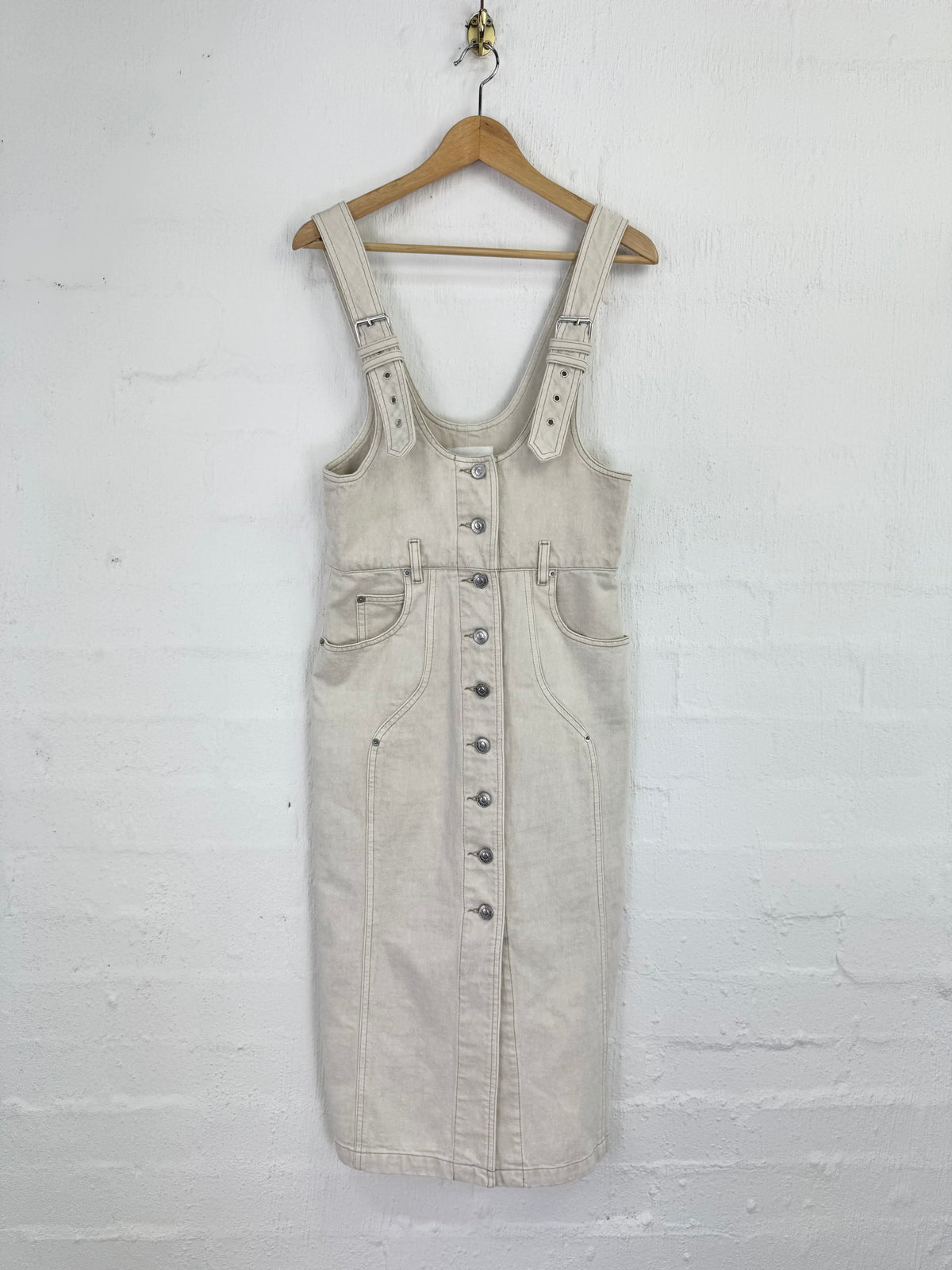 A light beige denim overall dress with front button closure and side pockets, hanging on a wooden hanger against a white painted brick wall.