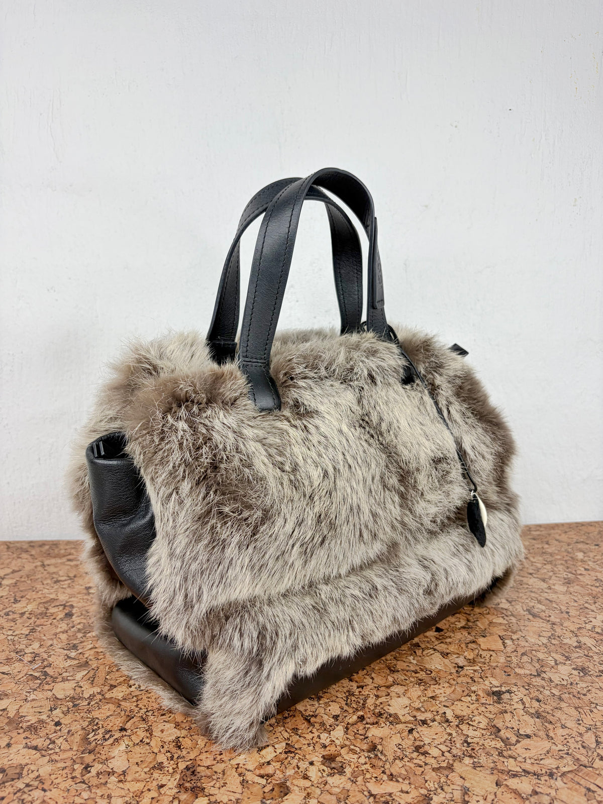 A gray and brown fur handbag with black leather handles and trim, resting on a brown speckled surface against a white wall.