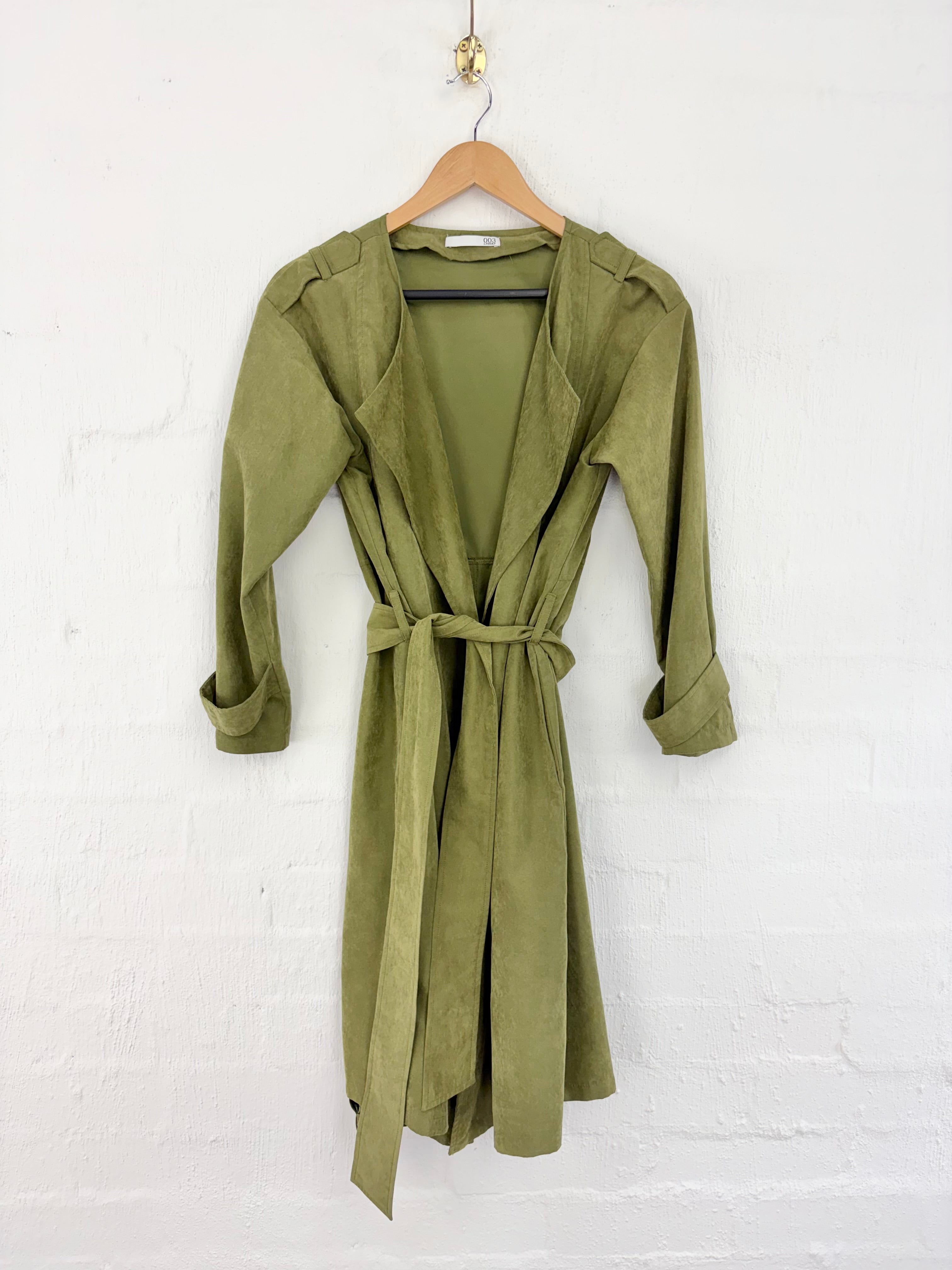 An olive green wrap dress with three-quarter sleeves and a matching waist tie, hanging on a wooden hanger against a white painted brick wall.