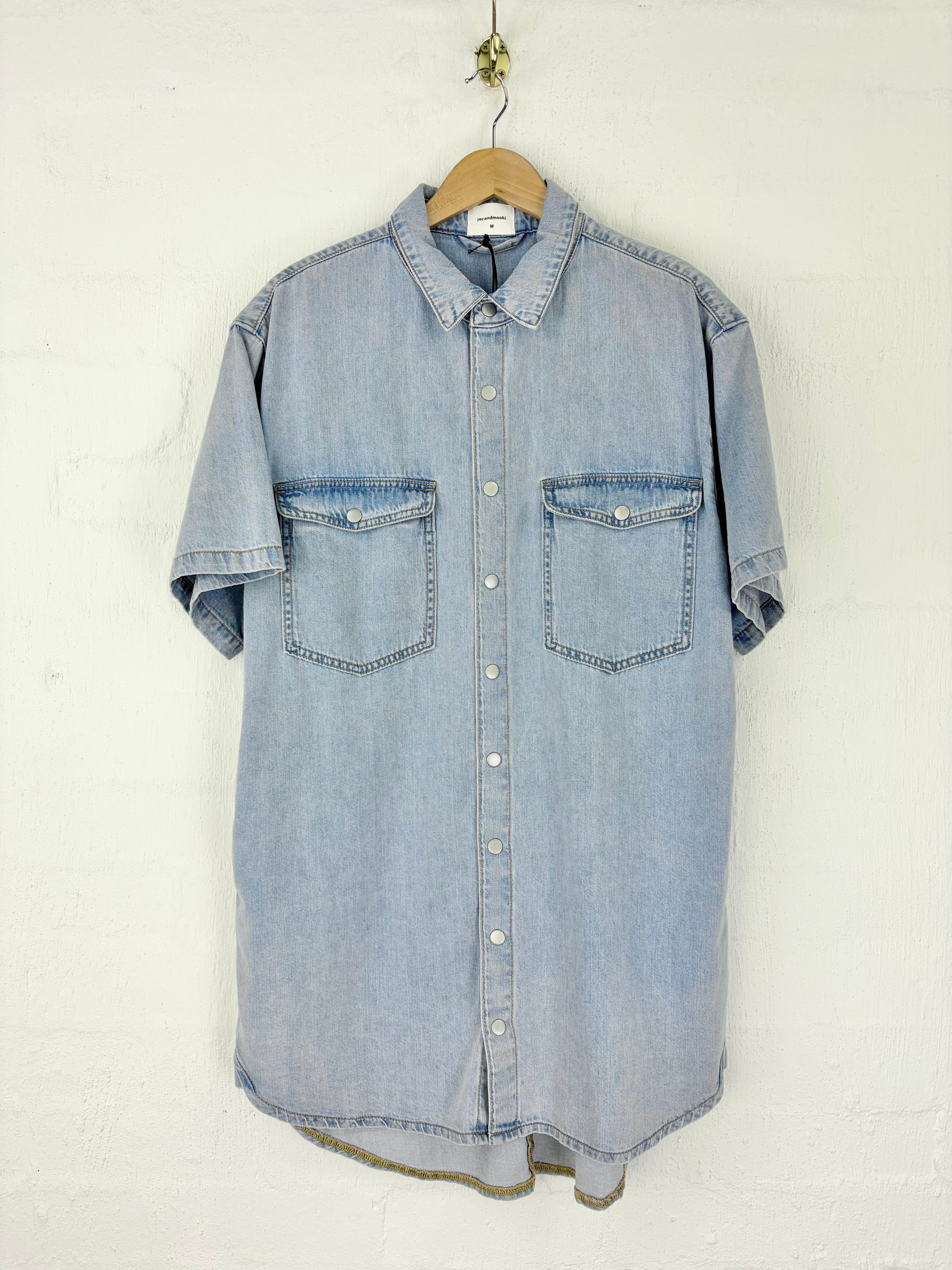 A light blue denim short-sleeve button-up shirt with two chest pockets, hanging on a wooden hanger against a white textured wall.