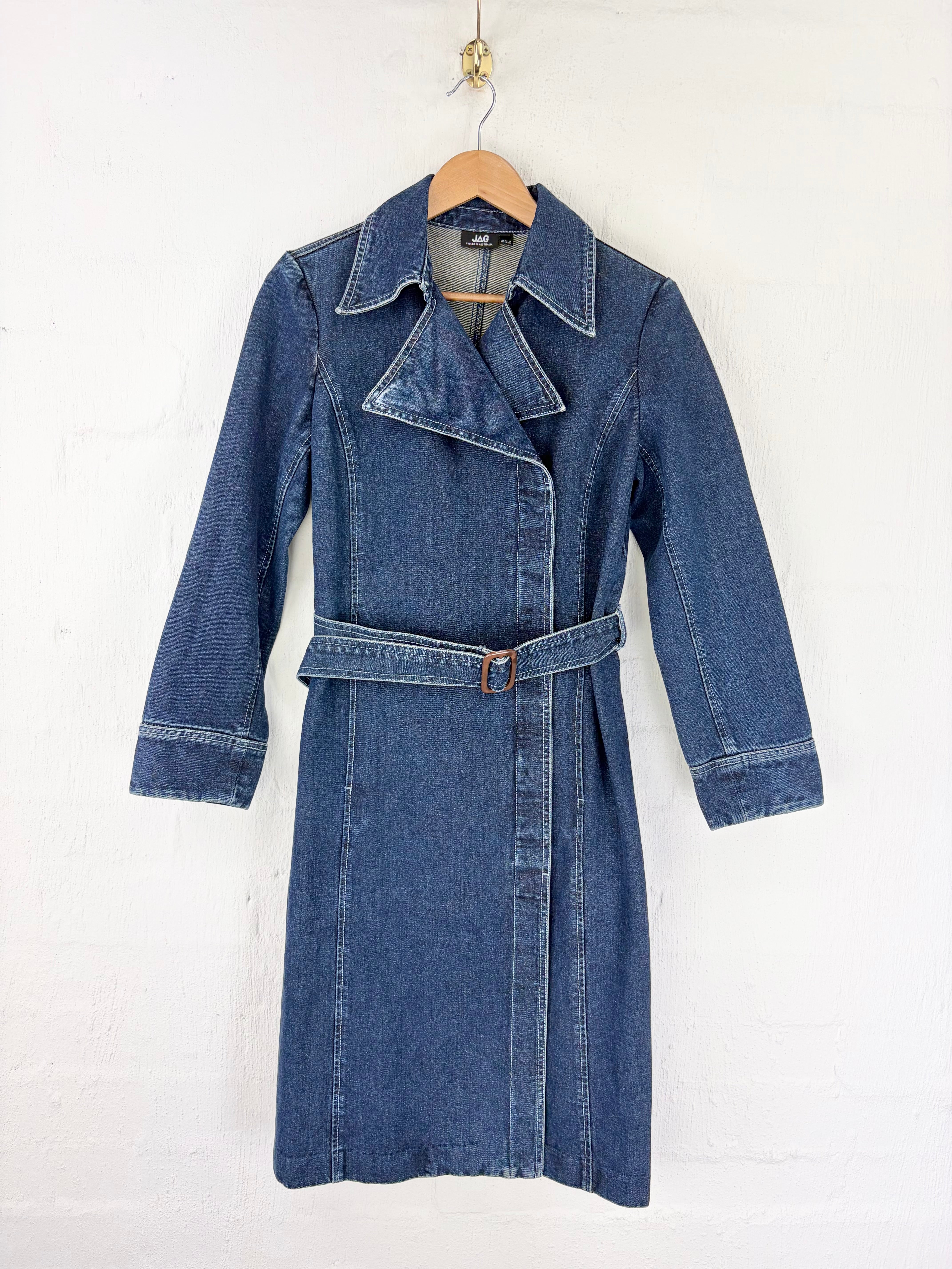 A blue denim trench coat with a matching waist belt, hanging on a wooden hanger against a white painted brick wall.