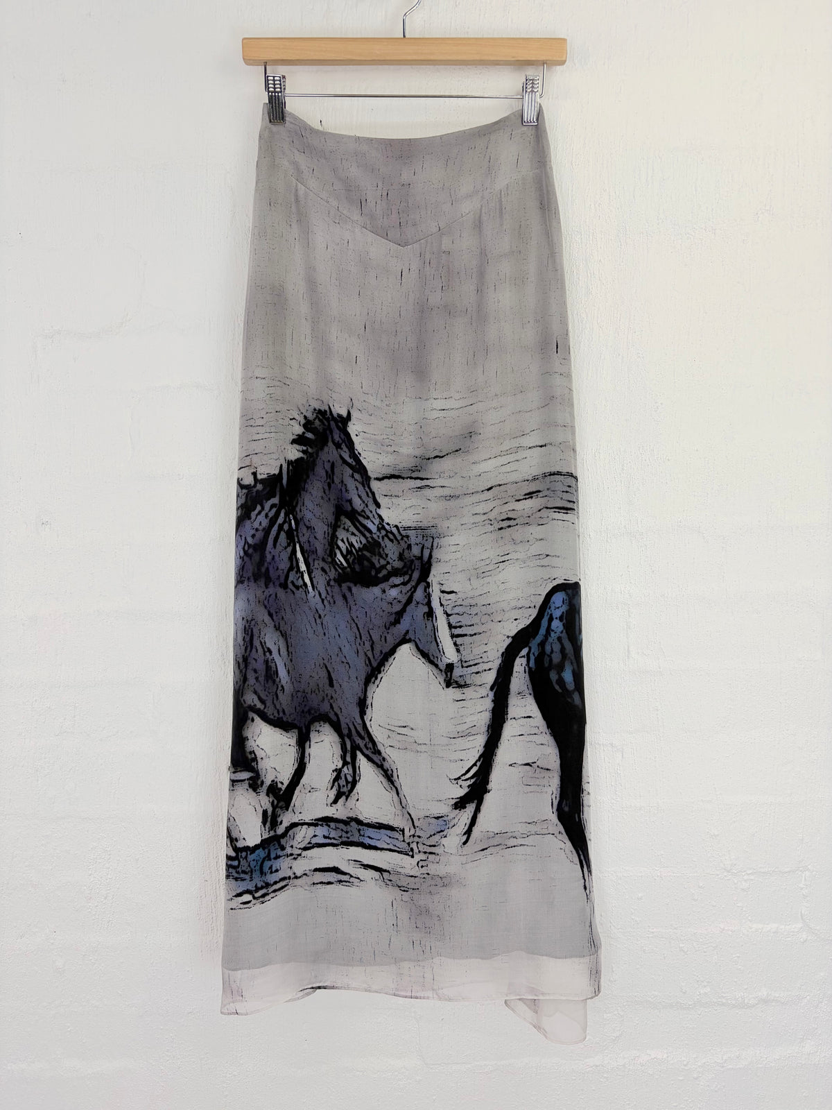 A long gray silk-like midi skirt featuring a black and blue painted horse design, hanging from a wooden hanger against a white painted brick wall.