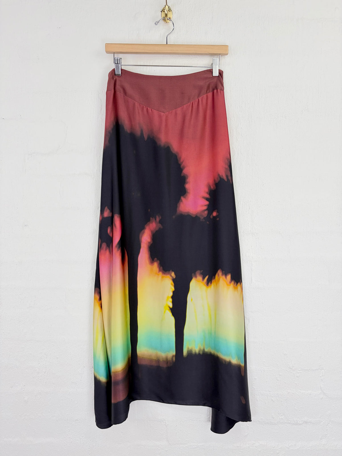 A midi skirt with a tie-dye sunset print, hanging on a wooden hanger against a white brick wall. The skirt features a dark base with a gradient of pink, red, yellow, green, and blue.