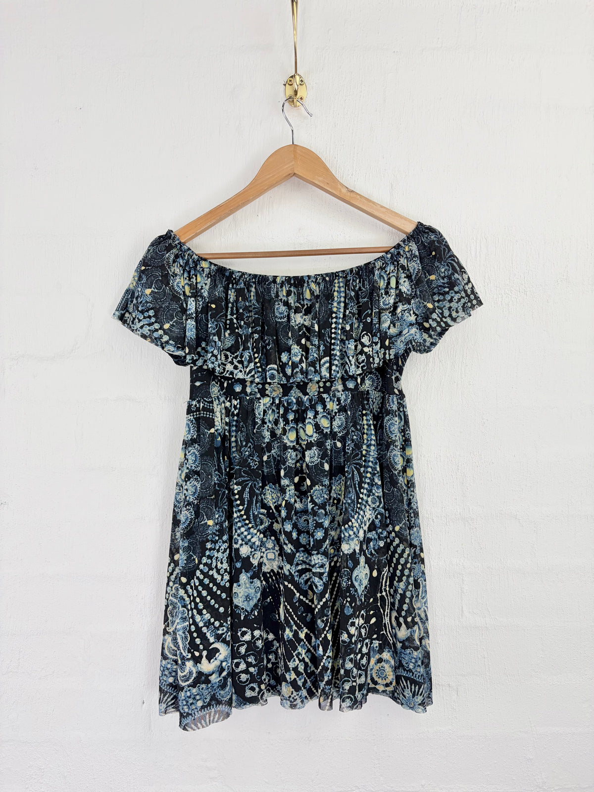 An off-the-shoulder short-sleeve dress with a blue, black, and white abstract floral pattern, hanging on a wooden hanger against a white brick wall.